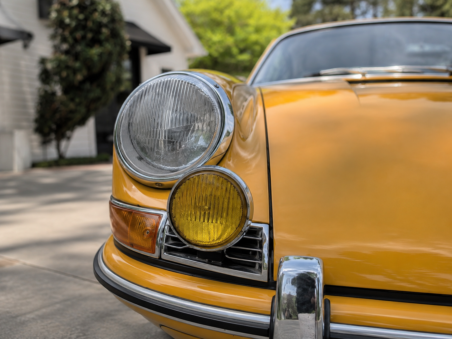Close-up of the front left side of a vintage yellow car showing the headlight, yellow fog light, and part of the bumper, with a house and trees in the background.