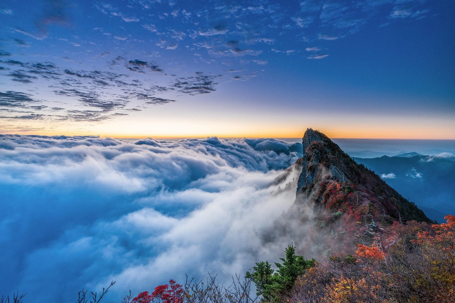 Sunrise view from a mountain peak with a cloud layer below, a rocky mountain ridge on the right, and a colorful sky with scattered clouds.