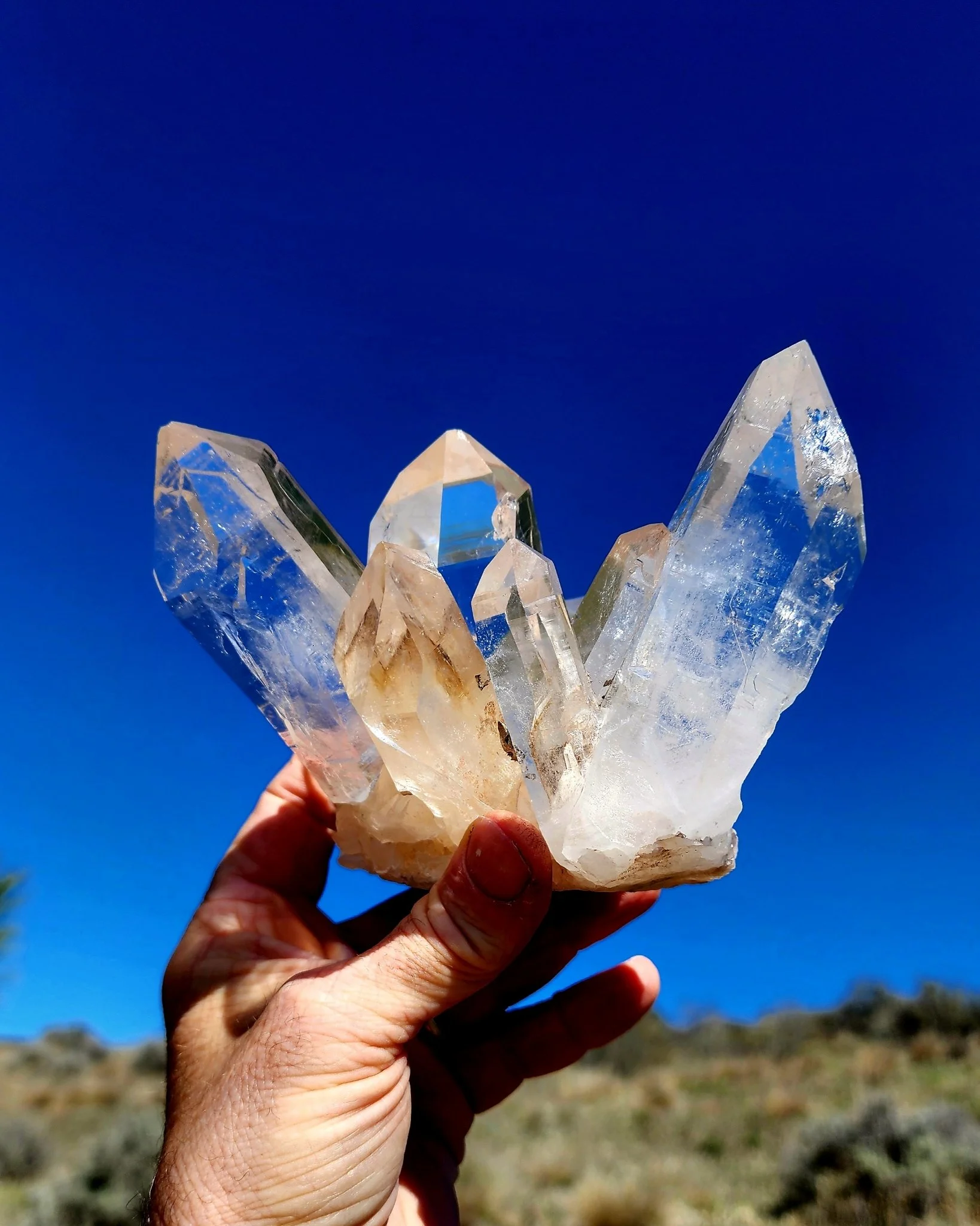 A person holding a cluster of clear quartz crystals against a bright blue sky. Large Himalayan Quartz Cluster. Mined at 18,000 feet in elevation in Himachal. Pradesh, India.