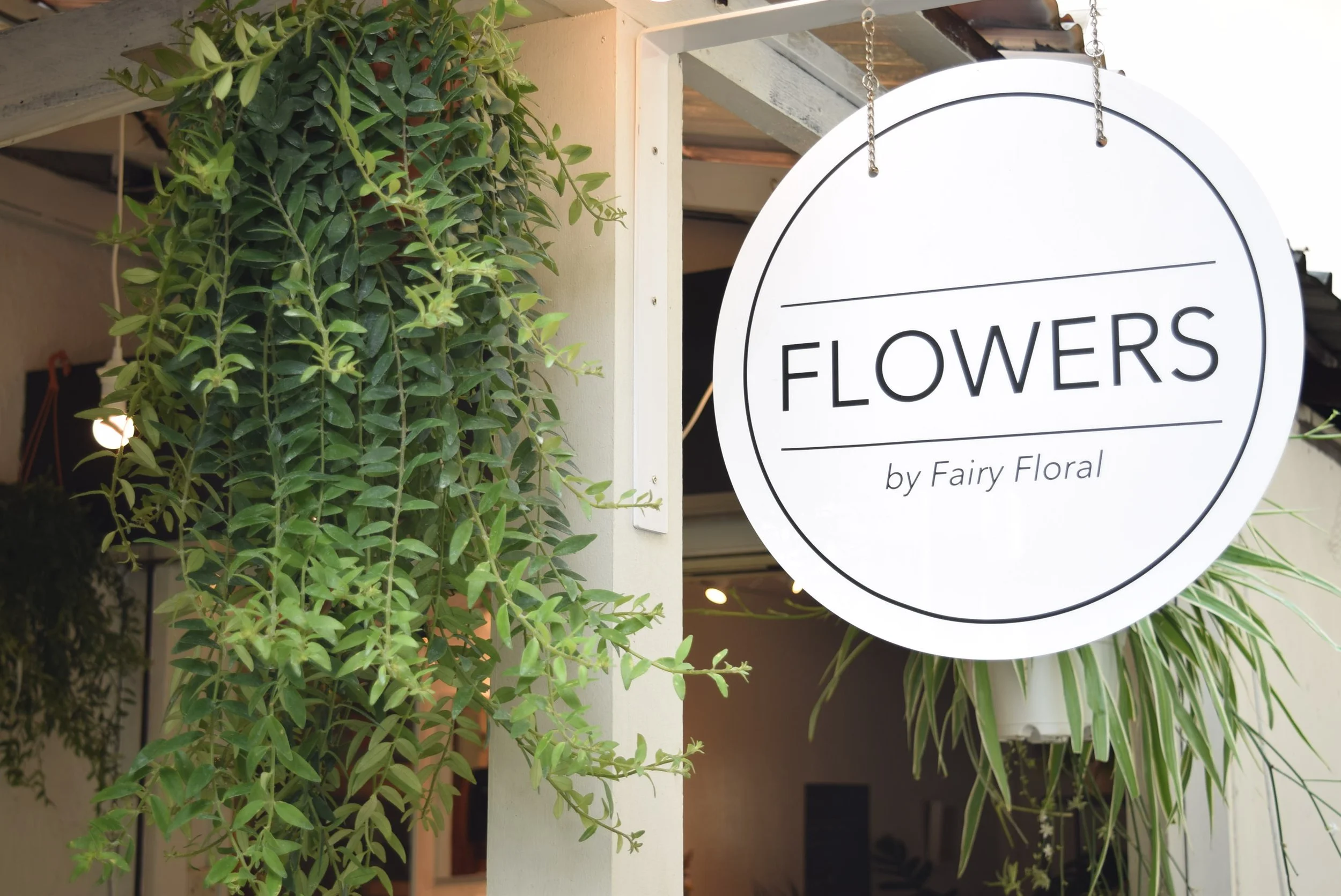 A circular sign hanging from a chain reads 'FLOWERS by Fairy Floral', mounted on a white frame. Green leafy plants surround the sign, with some visible in the background.