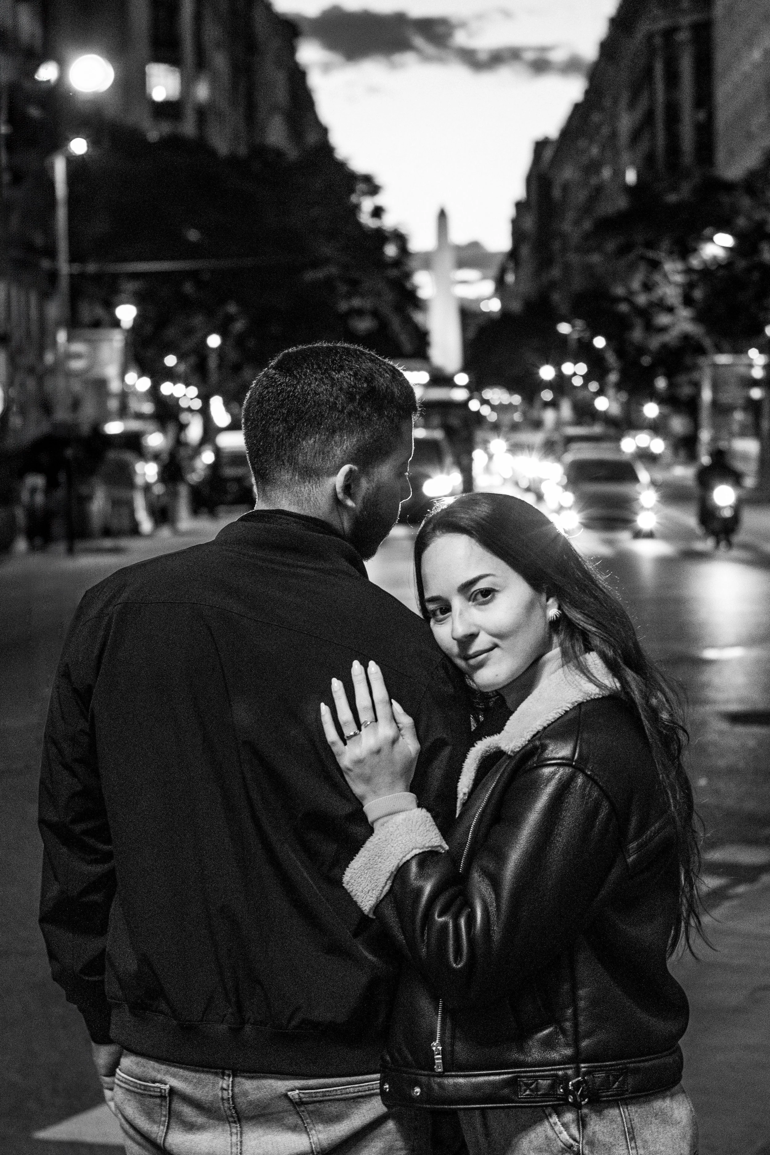 Pareja abrazada en una calle urbana de noche, en la imagen en blanco y negro. La mujer mira hacia la cámara y el hombre tiene la cabeza baja, con edificios y tráfico iluminado al fondo.