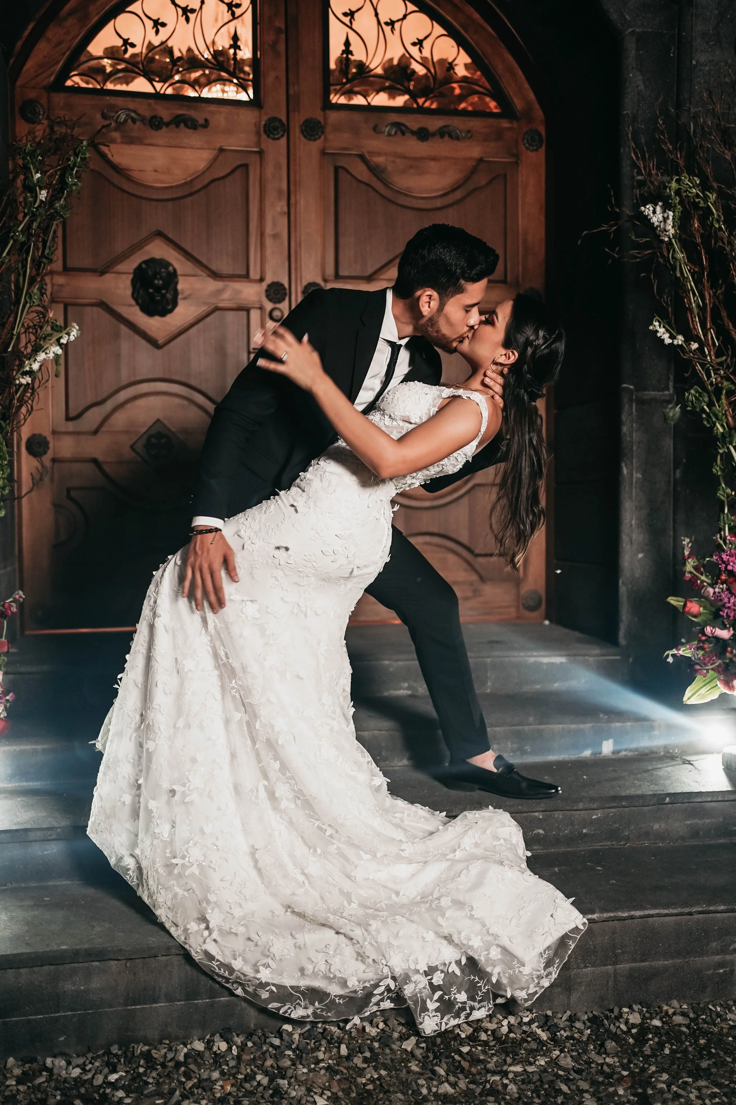 Una pareja de recién casados en un beso, ella con un vestido de novia largo y decorado con flores, él con traje negro, en la entrada de una casa de madera, rodeados de flores y plantas.