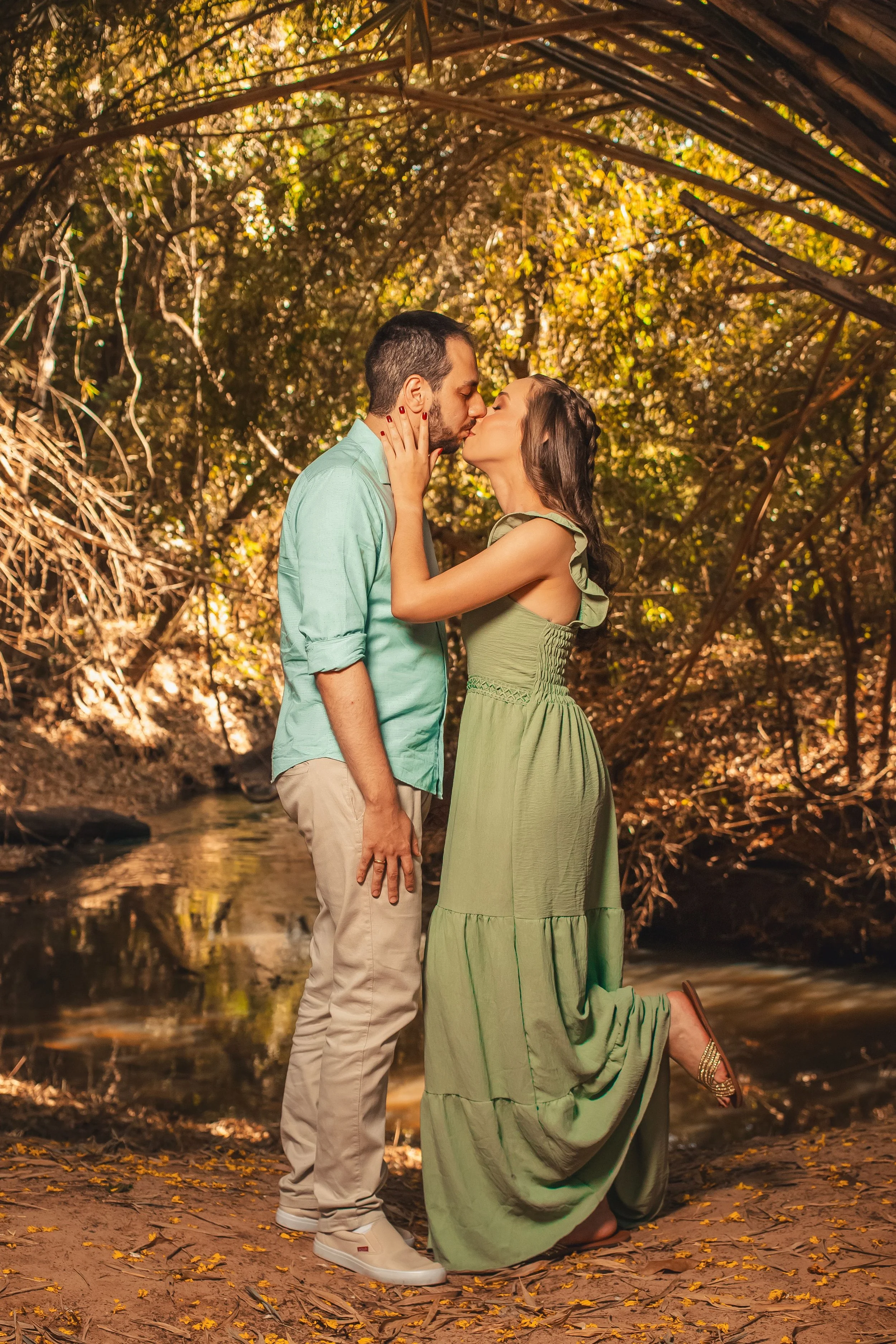 Una pareja en un bosque, la mujer está arrodillada y el hombre de pie, compartiendo un beso en un entorno natural con árboles y hojas secas en el suelo.