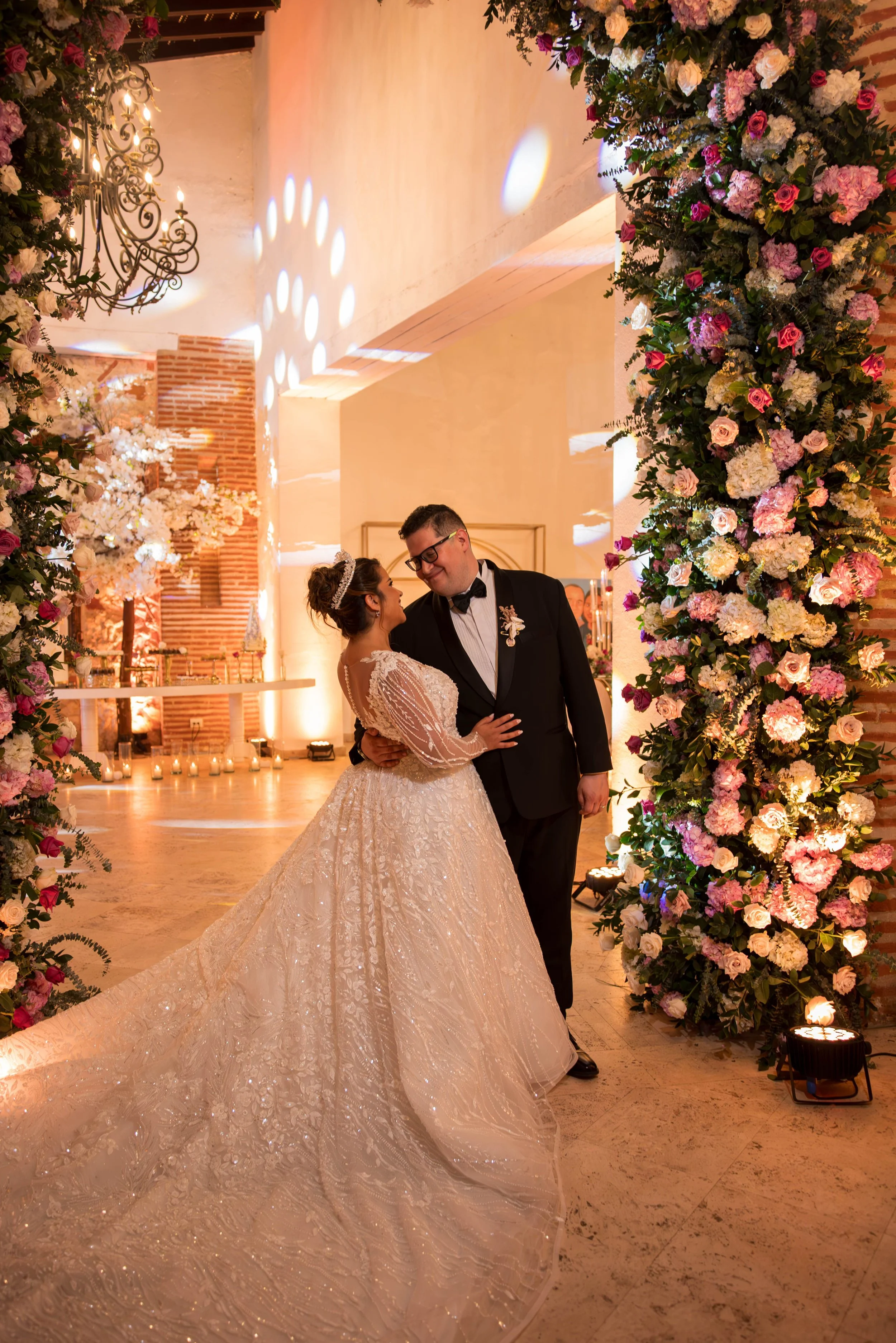 Pareja de recién casados en boda, la novia lleva vestido largo y brillante, el novio lleva esmoquin. Están sonriendo y mirándose en un arco decorado con flores rosas y blancas, en un lugar con paredes de ladrillo y decoraciones elegantes.
