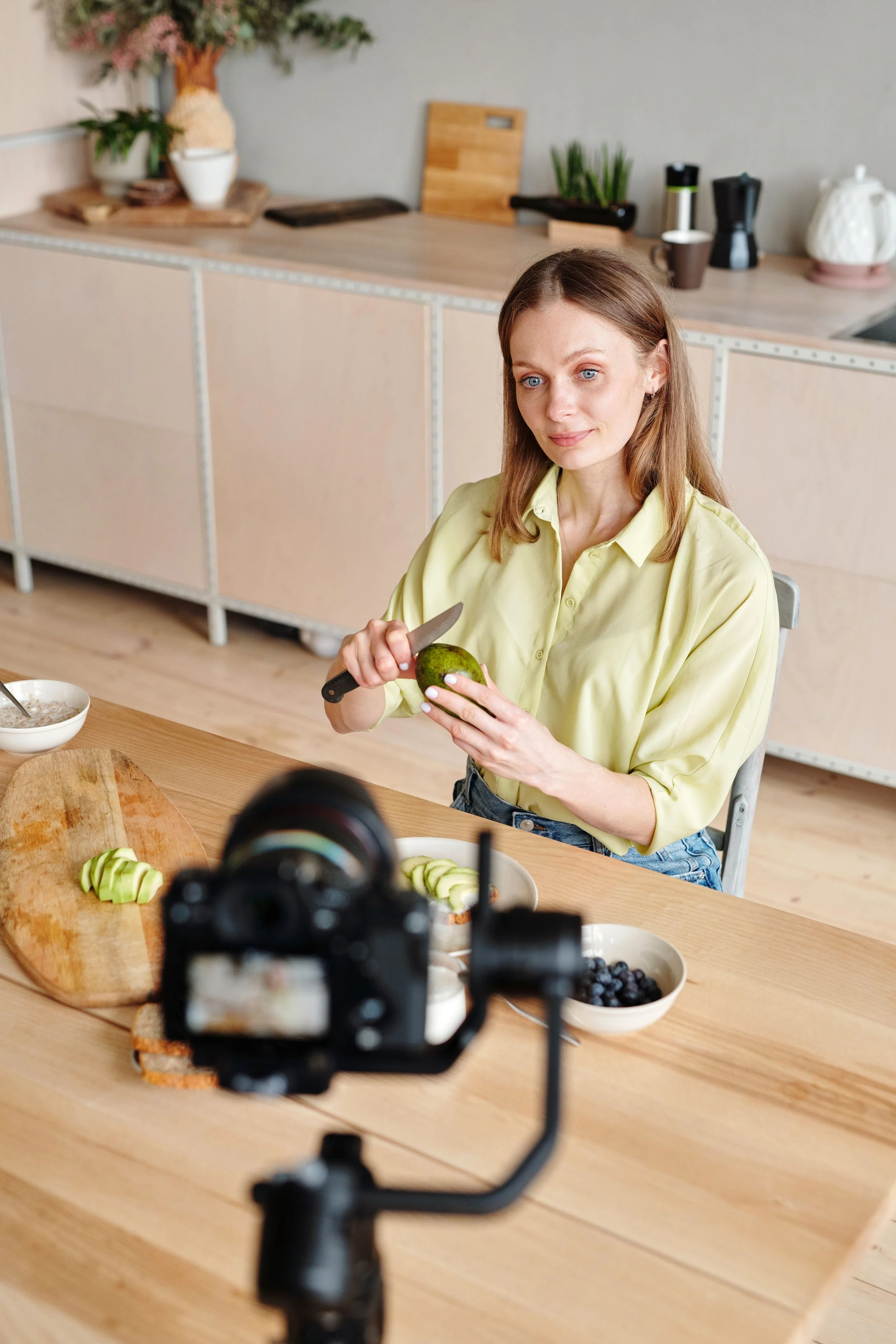 Mujer con cabello castaño claro y ojos azules, usando blusa amarilla, cortando un aguacate en una cocina moderna. En la mesa hay platos con fruta y cereal, y la cámara está grabando.