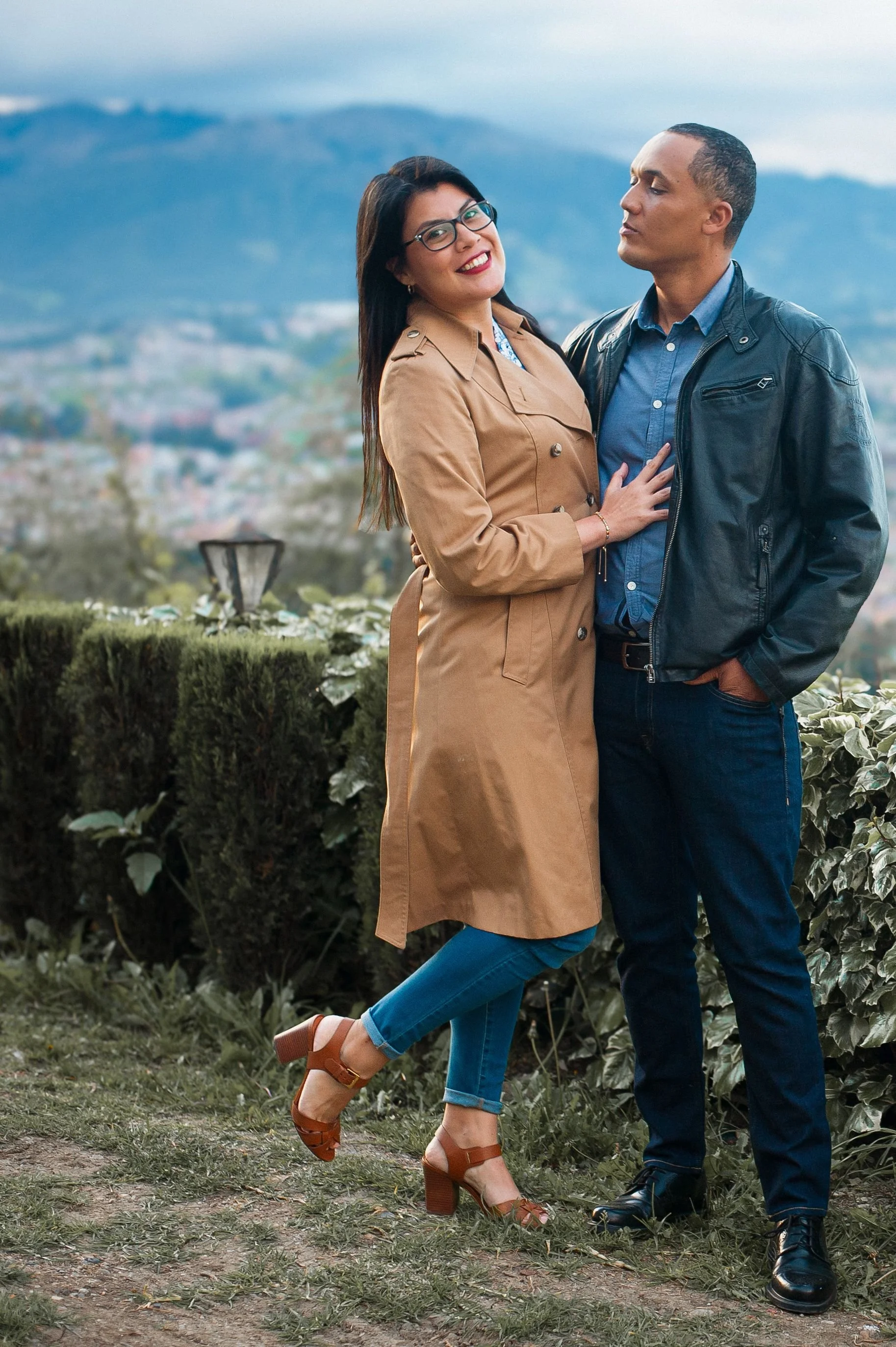 Una pareja posa juntos en un parque con fondo de montañas y cielo nublado. La mujer lleva gafas, un abrigo beige, pantalones azul claro y sandalias de tacón marrón. El hombre usa chaqueta de cuero negro, camisa azul y pantalones oscuros.