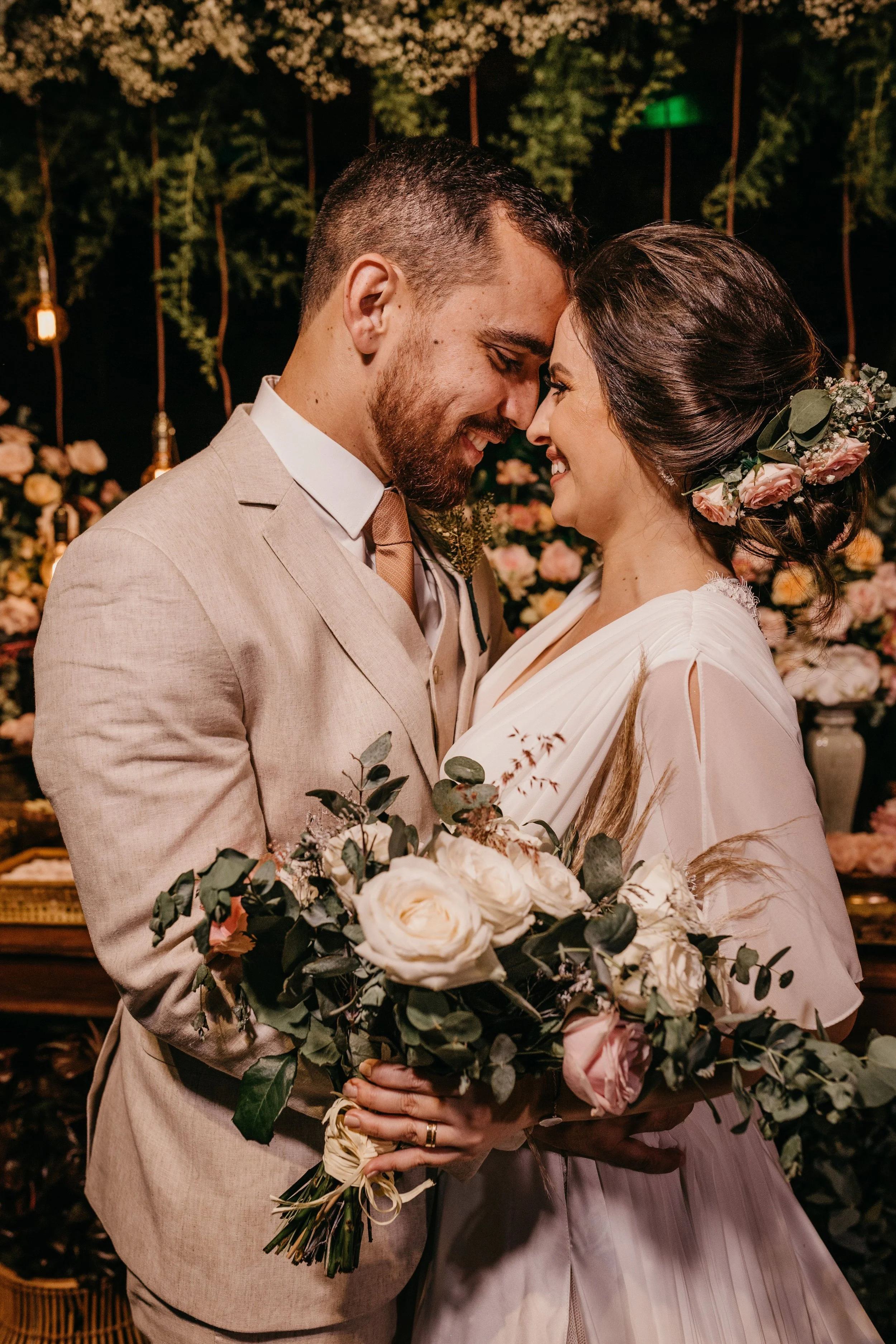 Una pareja de recién casados abrazados, con la frente tocándose, en una ceremonia nupcial con decoraciones florales y luces en el fondo.