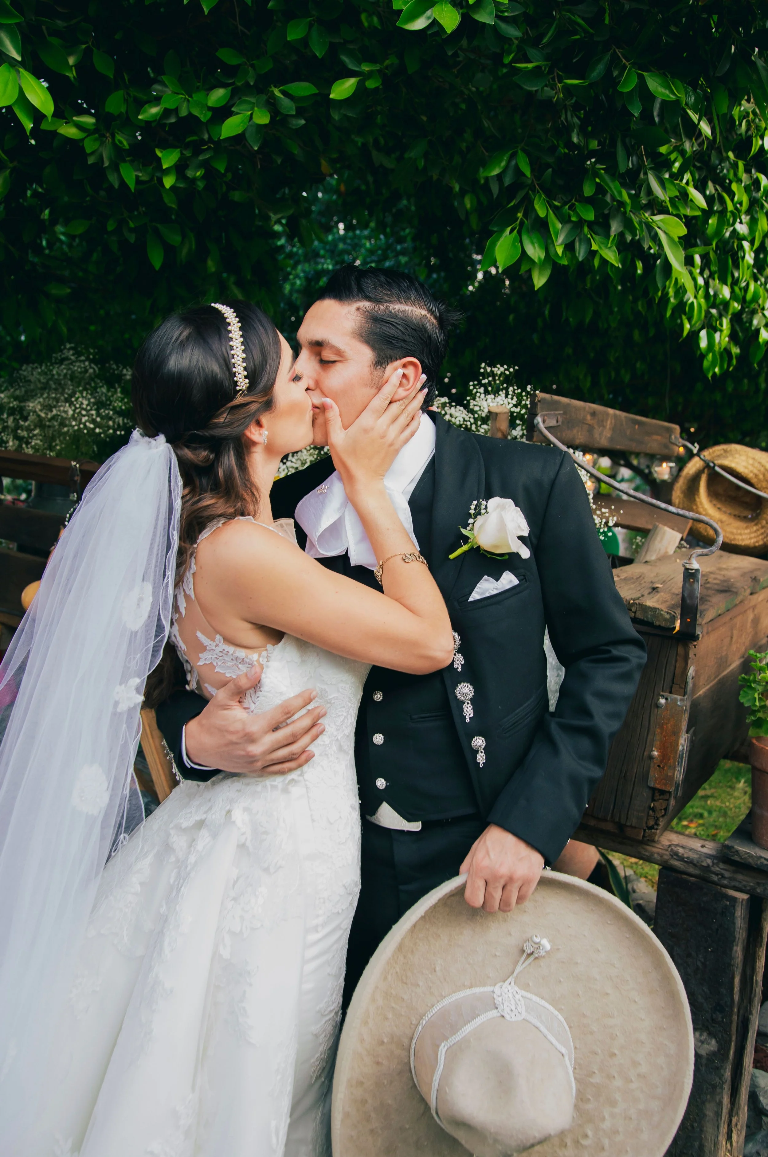 Pareja de novios en boda, la novia con vestido blanco y velo, el novio con traje negro, en un entorno natural con árboles verdes, en un momento de besarse.