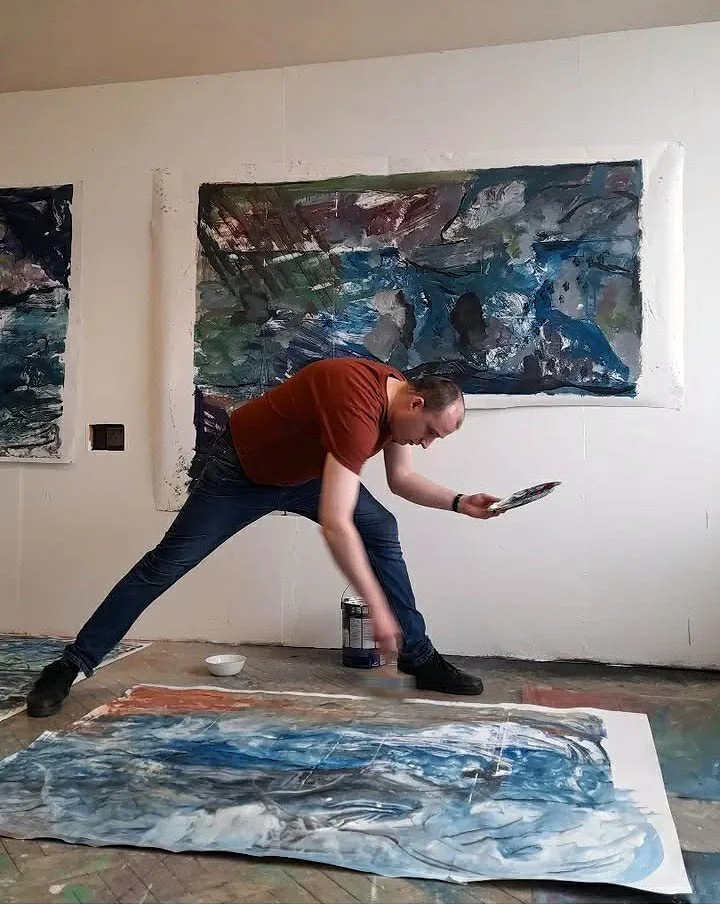 A man in a brown t-shirt and blue jeans standing in an art studio, holding a paint palette and reaching down towards a large, abstract watercolor painting on the floor, with another similar artwork hanging on the wall behind him.
