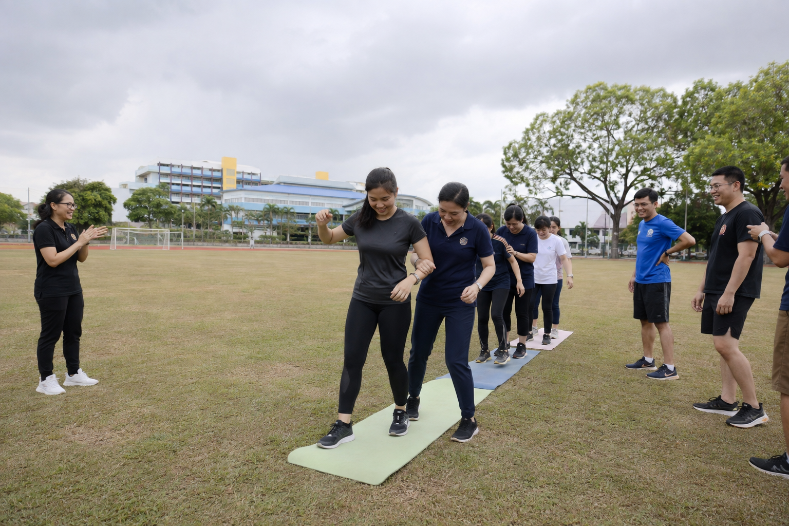 A group of people participating in an outdoor team-building activity on a grassy field, with some walking in a line on a narrow mat while others watch and cheer, under cloudy skies.