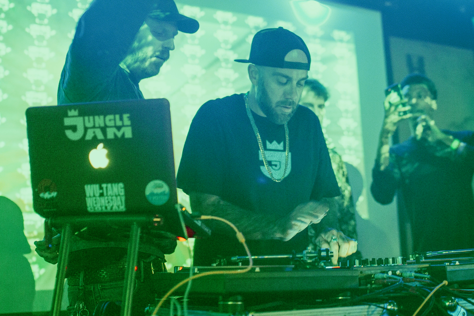 Two DJs performing at an event, with one of them looking at a phone. The laptop has stickers including one that says 'Jungle Jam' and an Apple logo. The background features a screen with repeating patterns and green lighting.