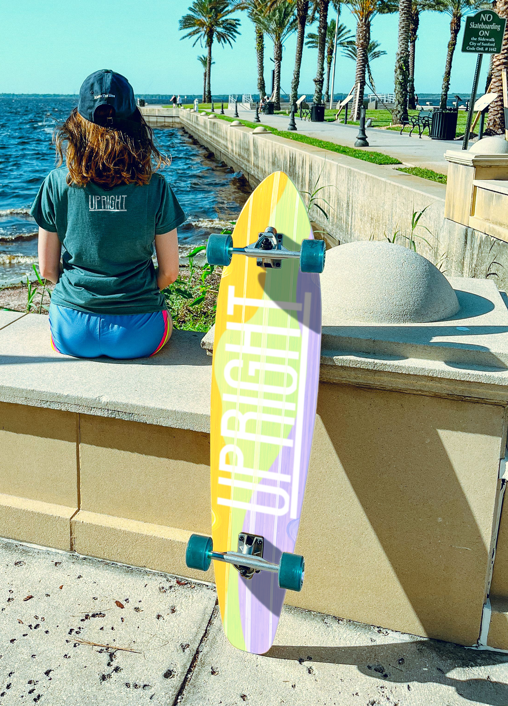 A person sitting by the water with a skateboard next to them, facing the ocean on a sunny day with palm trees along the promenade.