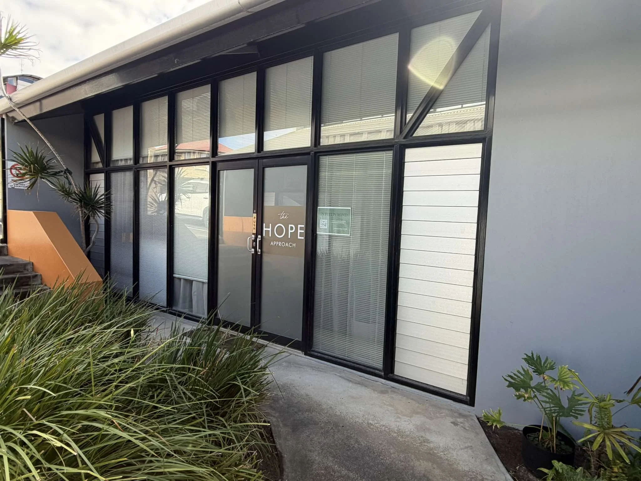 A modern building with large glass windows and a door labeled "the HOPE approach." There are plants and shrubs in front of the building and a concrete walkway.