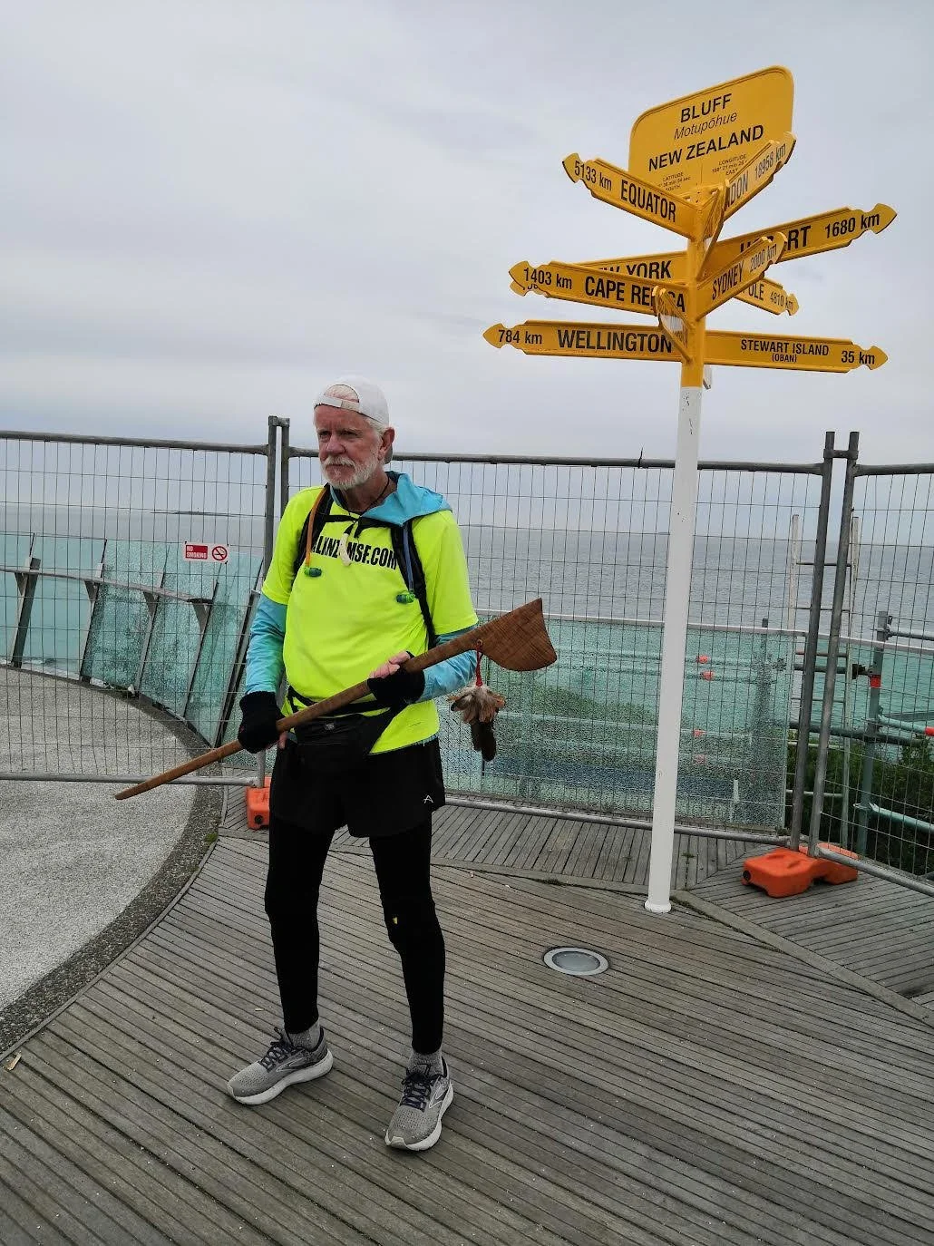 An elderly man holding a wooden paddle standing on a wooden deck near a street sign at a lookout point. The street sign points to various global destinations including New Zealand, Equator, Cape Reinga, Wellington, and Stewart Island. The man is wearing a bright yellow shirt, black leggings, and running shoes, and has a white beard and cap.