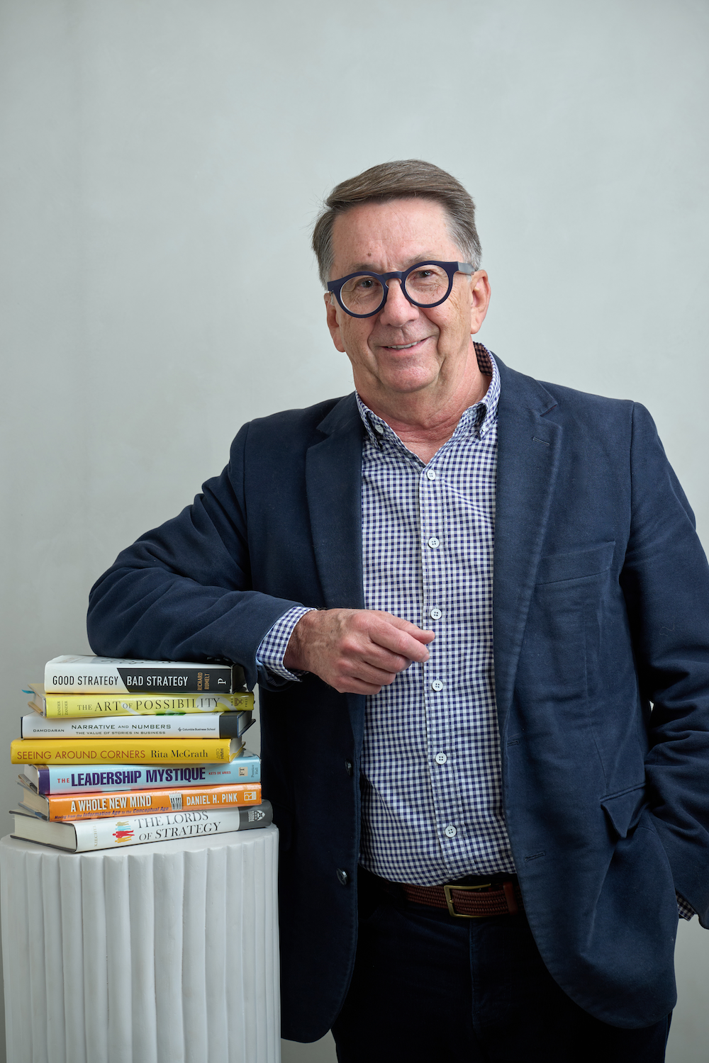 Dr David Blyth in a navy blazer and checkered shirt poses next to a stack of books on a white pedestal, smiling at the camera.