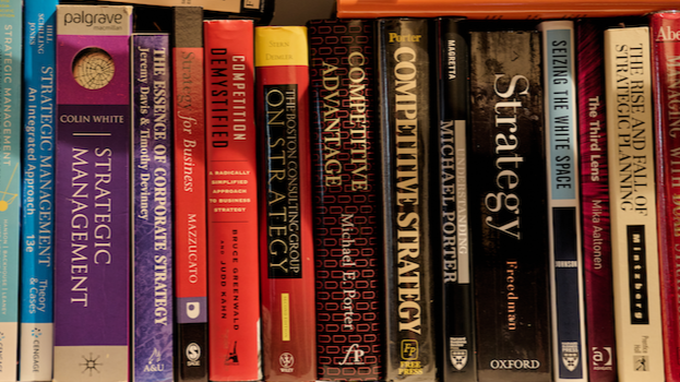 Books on a shelf about business strategy and management, including titles like 'Strategic Management,' 'Competitive Demystified,' 'Competing for Business,' 'Strategic Thinking,' 'Strategy,' and 'The Rise and Fall of Strategic Space.'