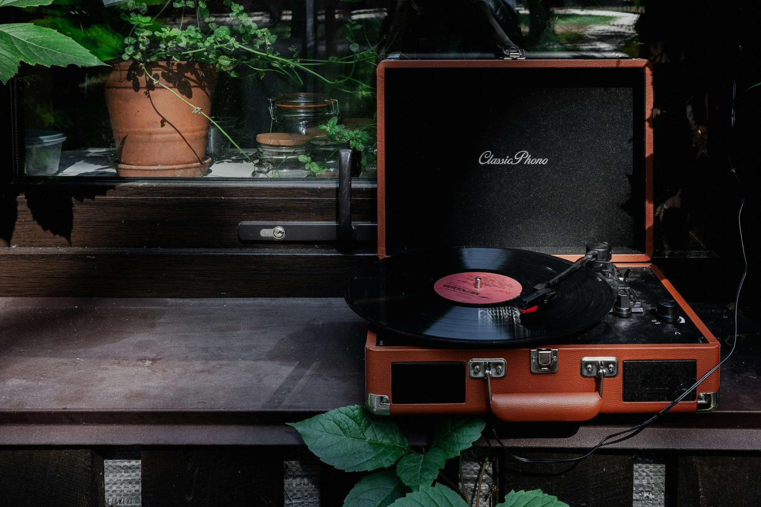 A vintage record player on a wooden surface along with a speaker labeled 'ClassiaPhono.' There are plants in the background and a window with glass. The record player has a pink label on the record and the surroundings have natural lighting.