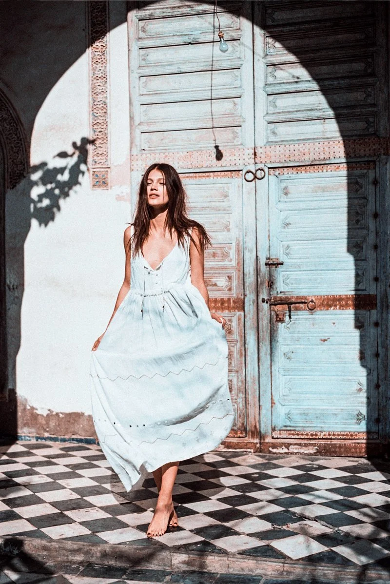 A woman in a white dress walking barefoot on a checkered floor in front of a weathered wooden door, with sunlight casting a shadow and a light bulb hanging from the ceiling