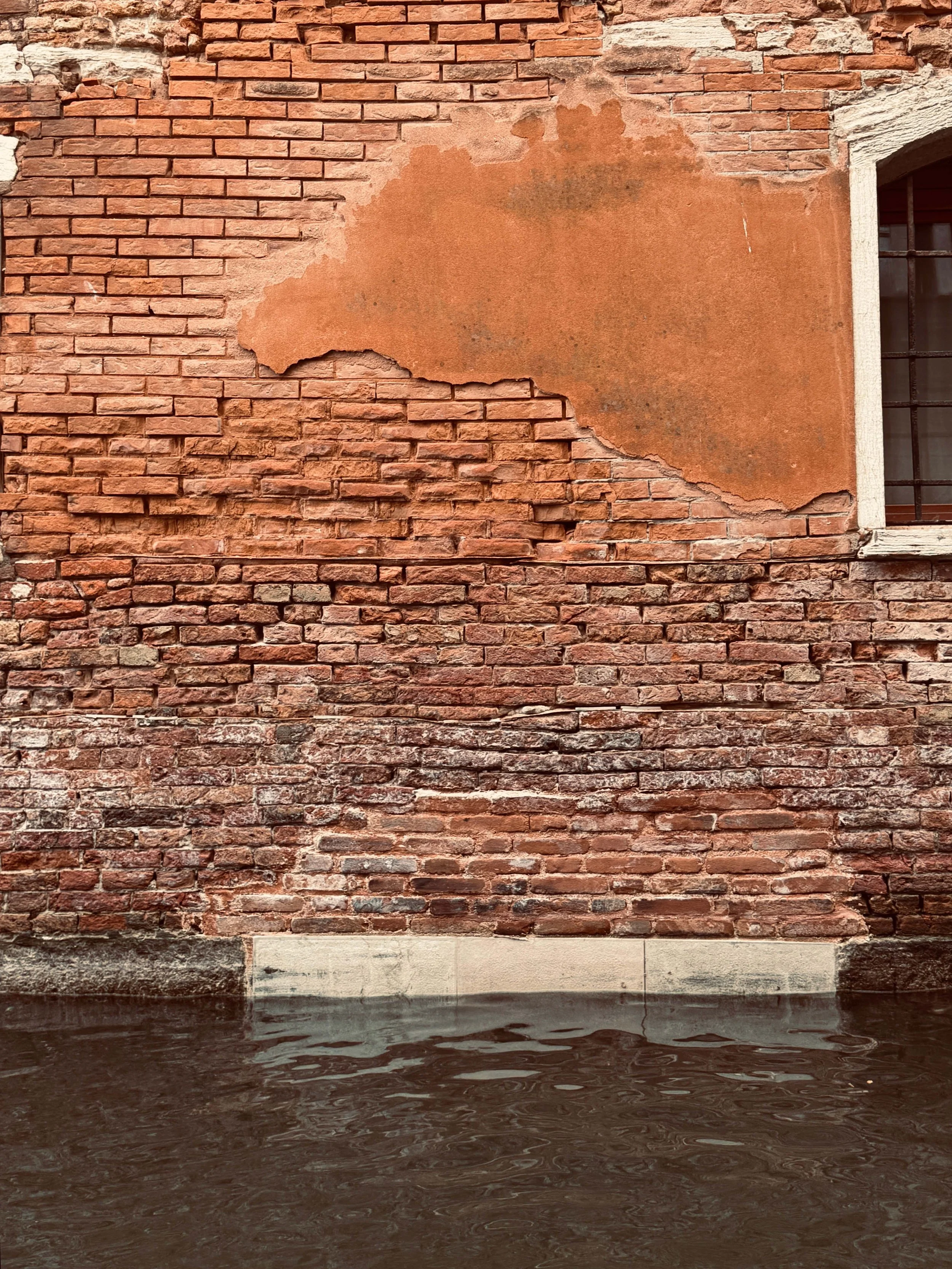 A brick wall with some missing and peeling sections, adjacent to a water-filled canal at the bottom.