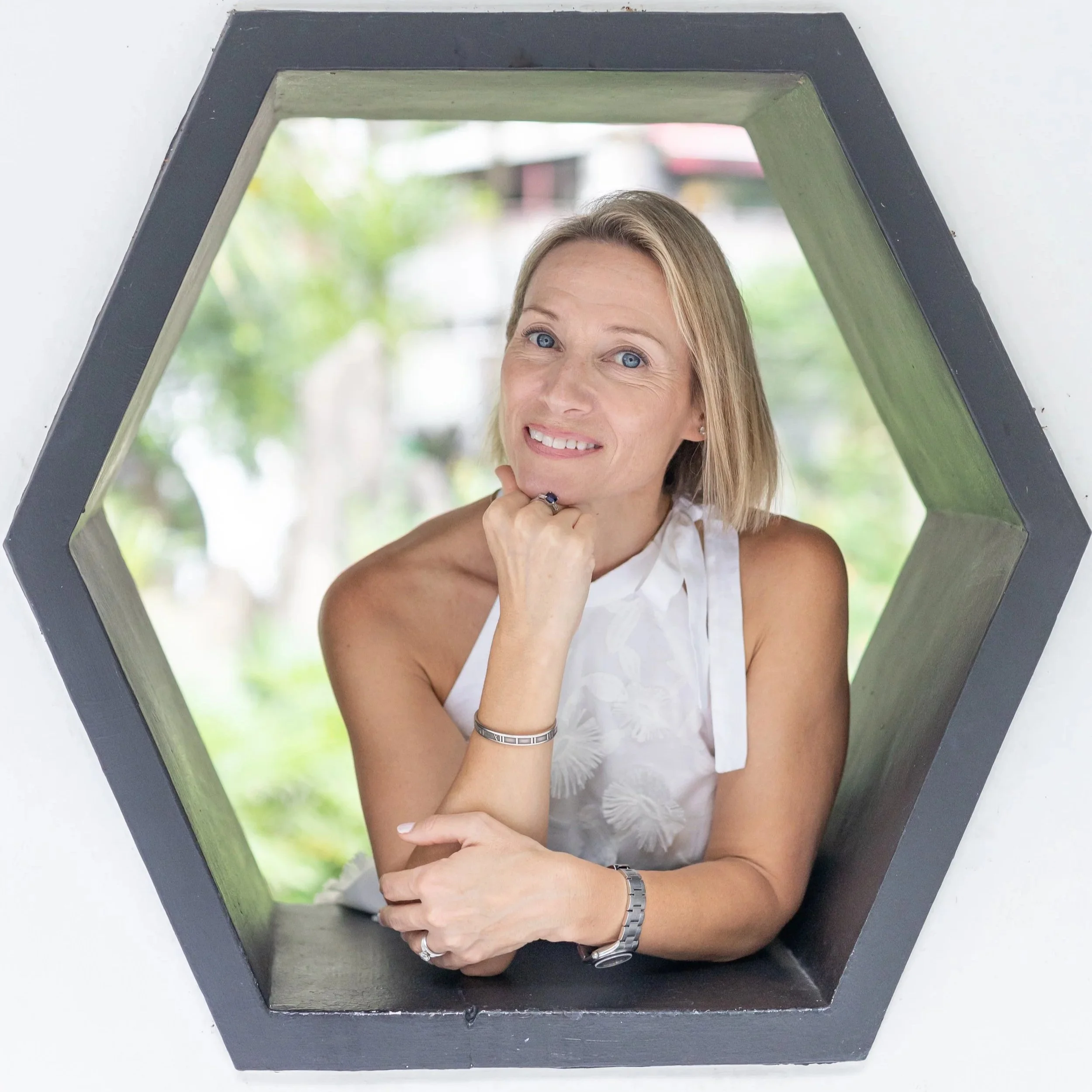 A woman with blue eyes and long blonde hair is looking in a mirror, smiling, and resting her chin on her hand. She is wearing a white sleeveless top and a silver wristwatch. The mirror has a black frame, and the background outside the mirror appears to be a blurred outdoor scene with greenery.