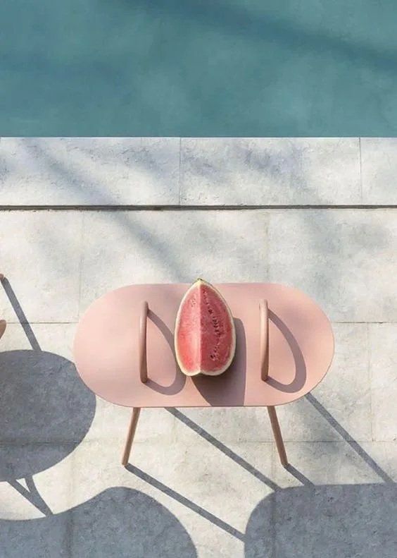 A pink outdoor chair with a slice of watermelon placed on its seat, casting a shadow on a tiled ground near a pool.