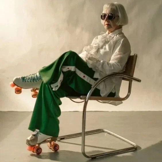 A person with white hair wearing sunglasses, a white blouse, and green pants with white and black stripes, sitting on a metal chair with their legs crossed on a skateboard, against a plain light-colored background.