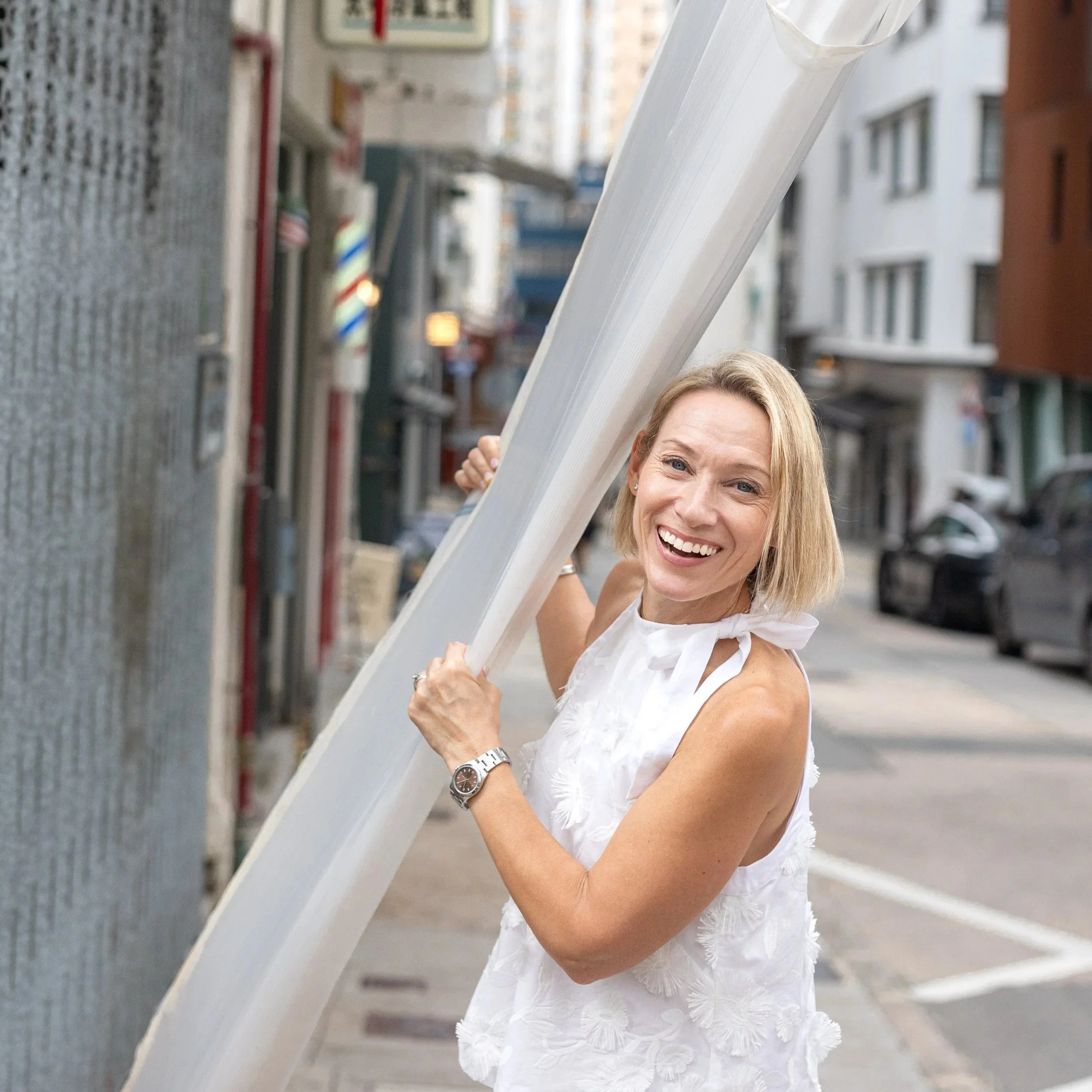 A woman with blonde hair, smiling, wearing a white sleeveless dress with floral patterns, holding a white rectangular object, standing on a city street with buildings and a barber pole in the background.