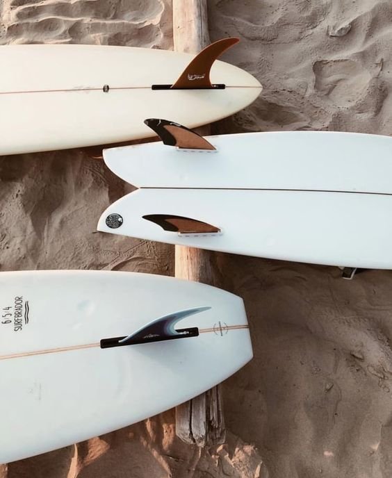Three surfboards leaning against a wooden post on sandy ground with a stone wall in the background.