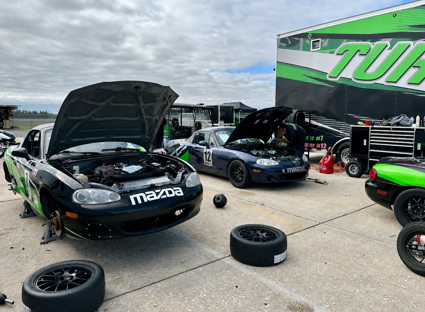 Race cars in a pit area with open hoods, mechanics working, and spare tires on the ground.