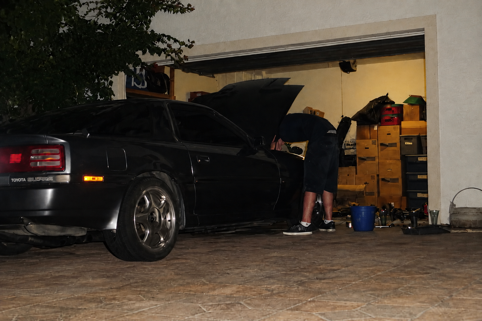 A man working on a gray Toyota Supra in front of a garage at night. The garage has various tools and storage shelves.