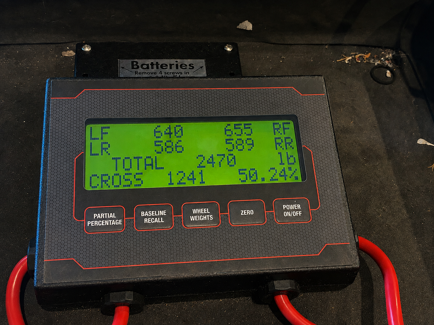 A digital tire balancing machine displaying wheel alignment data with readings for left front, left rear, right front, and right rear tires, along with total and cross weights, on a black work surface with some debris around.