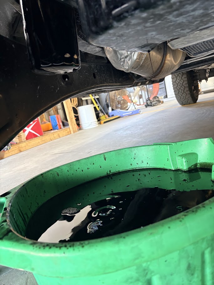 The image shows the underside of a vehicle in a garage, with a green container filled with dark oil or fluid positioned underneath to catch draining fluid during an oil change or maintenance.