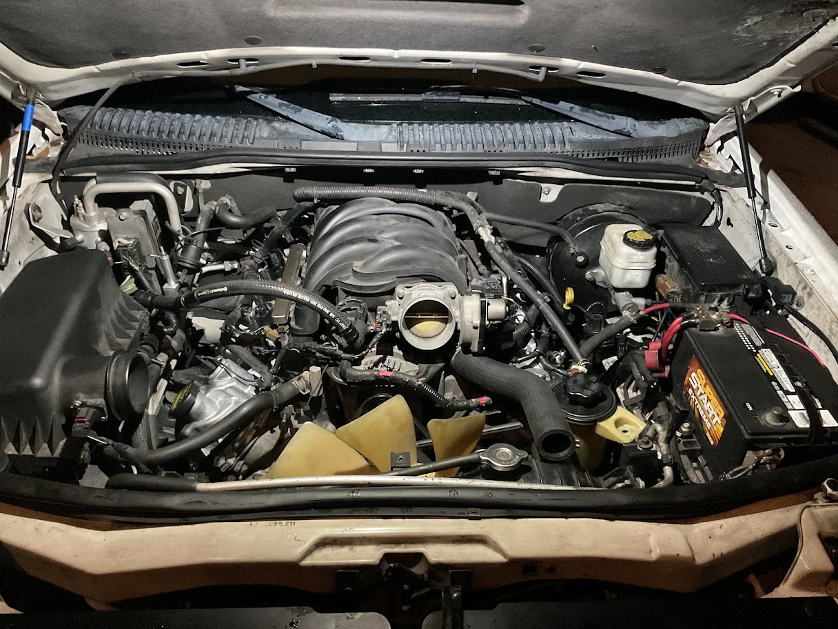 An open car hood showing the engine bay with various engine components, a battery, and fluid containers.