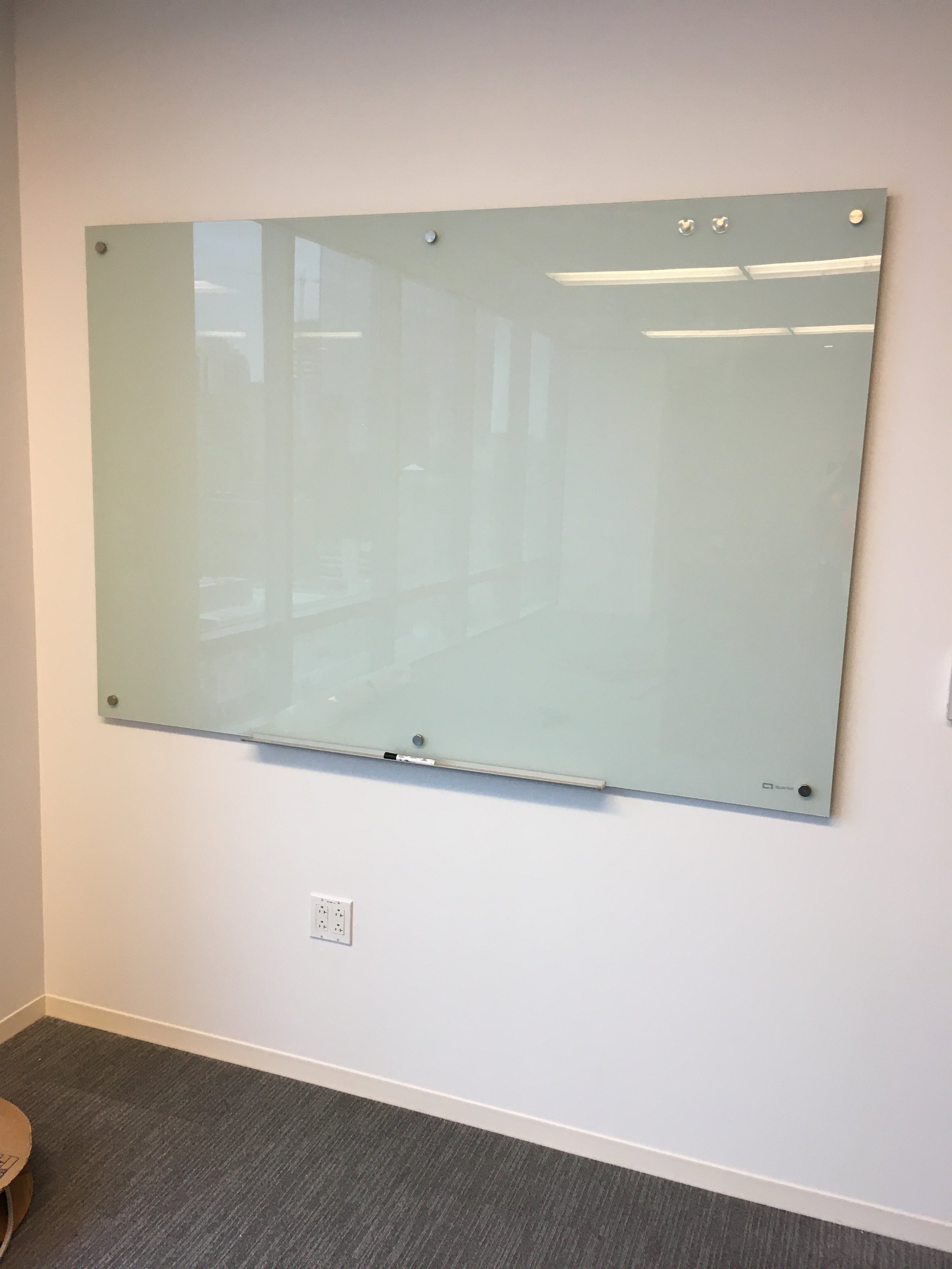 Empty whiteboard mounted on a wall in an office room with gray carpeted floor and electrical outlets below.