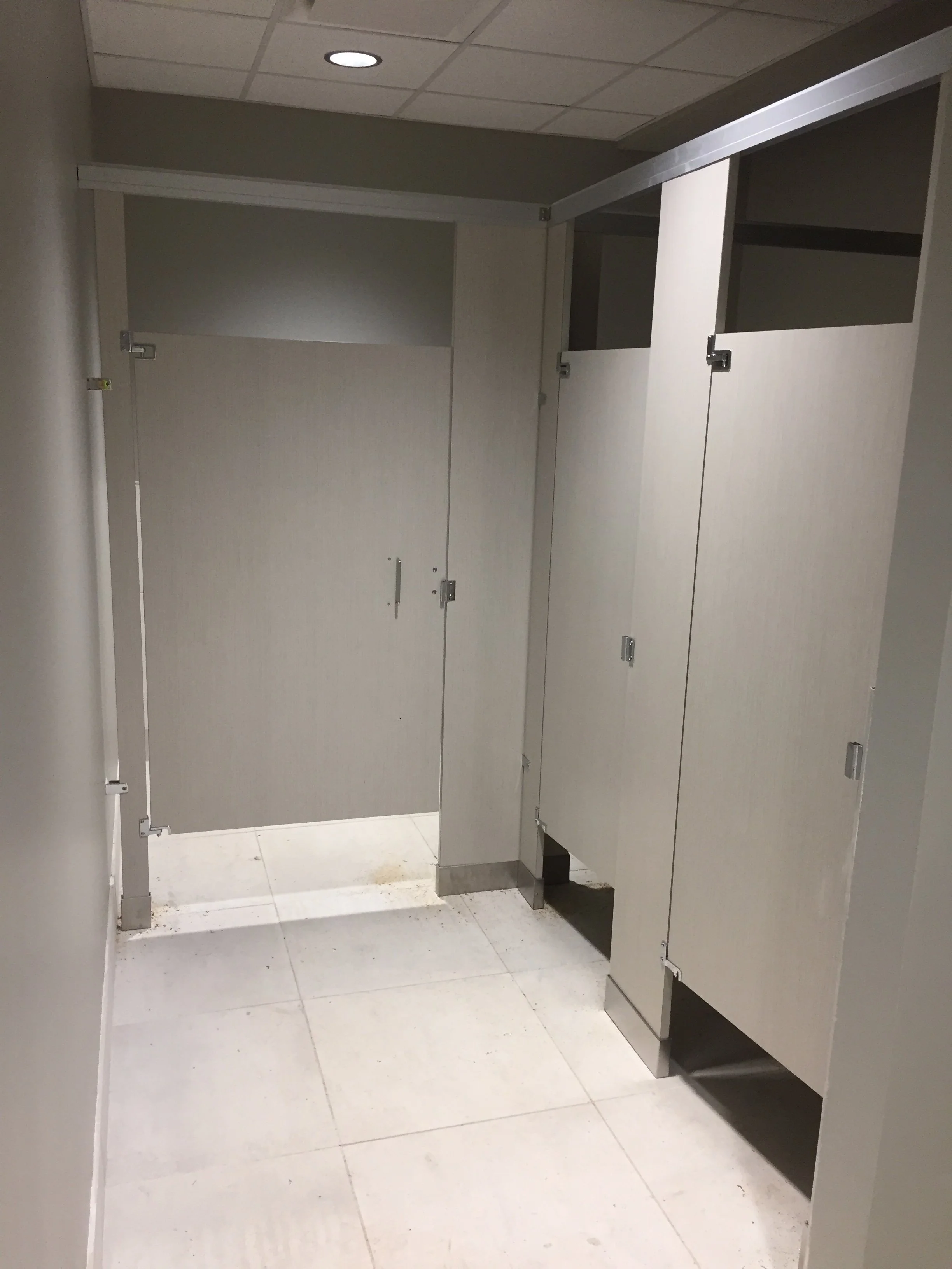 Public restroom with grey stalls, some open, in a tiled floor bathroom with a ceiling light.
