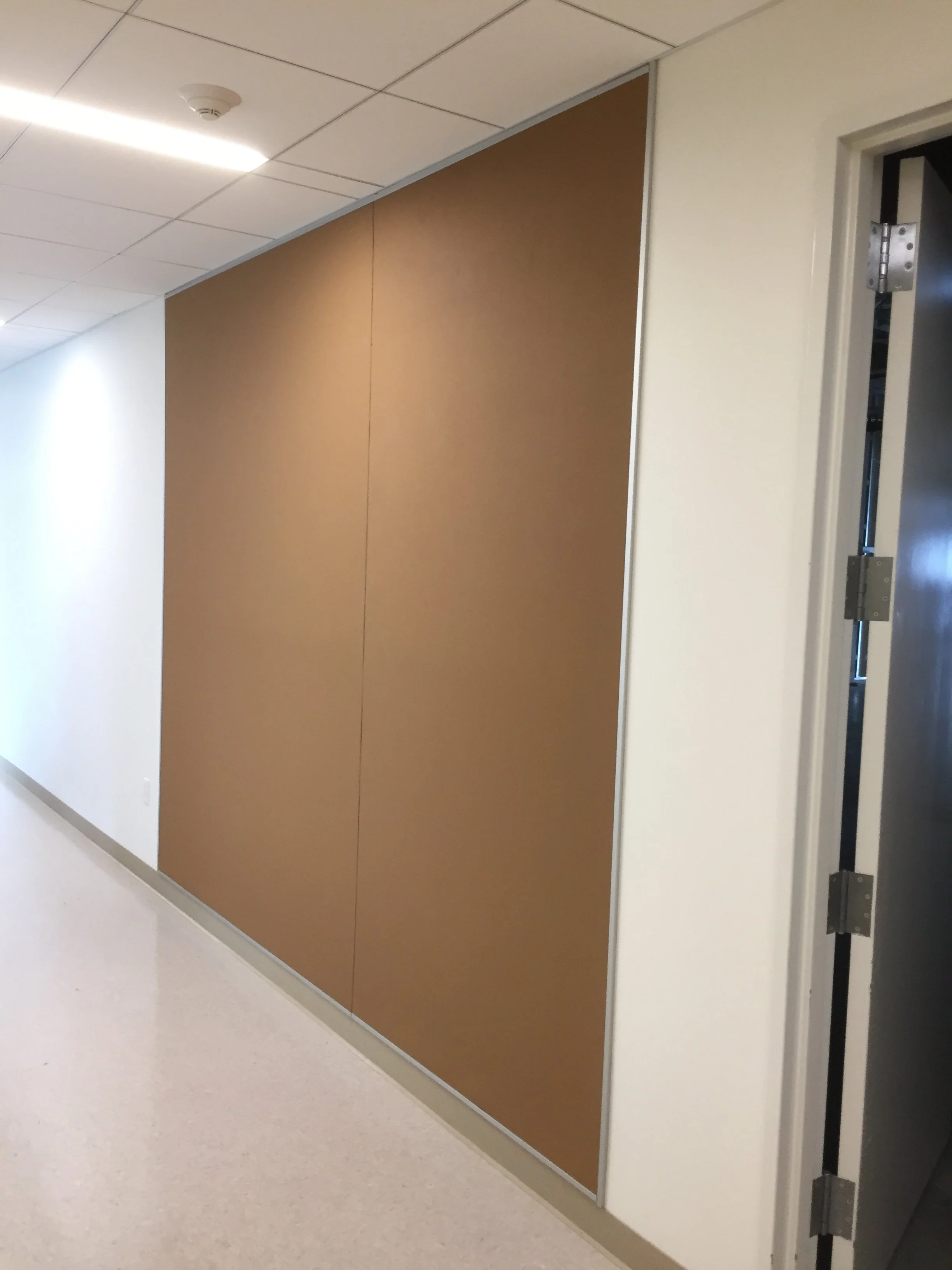 An indoor hallway with a ceiling light, smoke detector, and beige tiled floor. On the right side, there's a door open to an elevator with visible mechanical components inside. A large brown partition wall is on the left side of the hallway.
