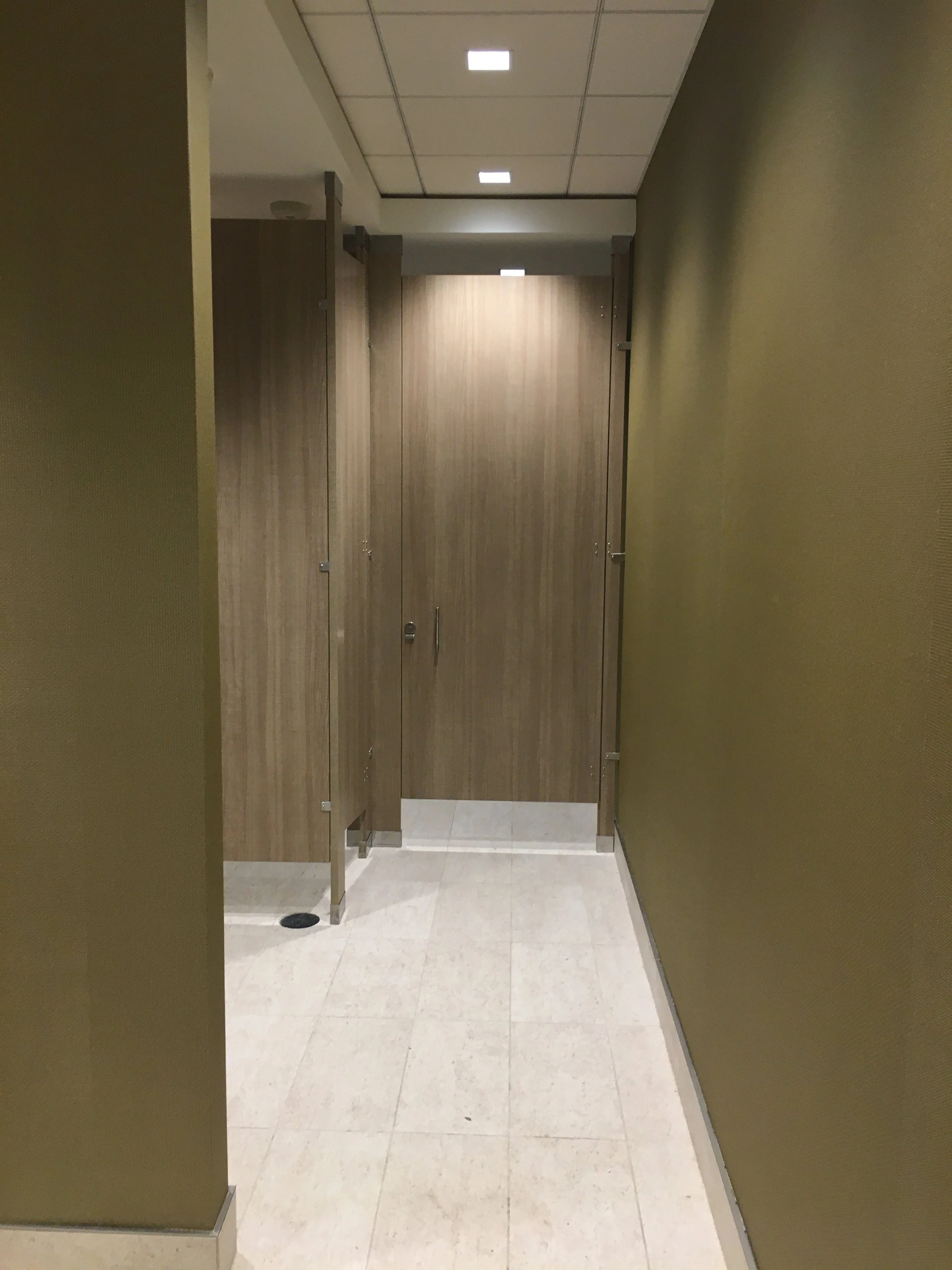 Image of a restroom stall area with beige tiled floor, wood grain partition doors, and olive green walls.