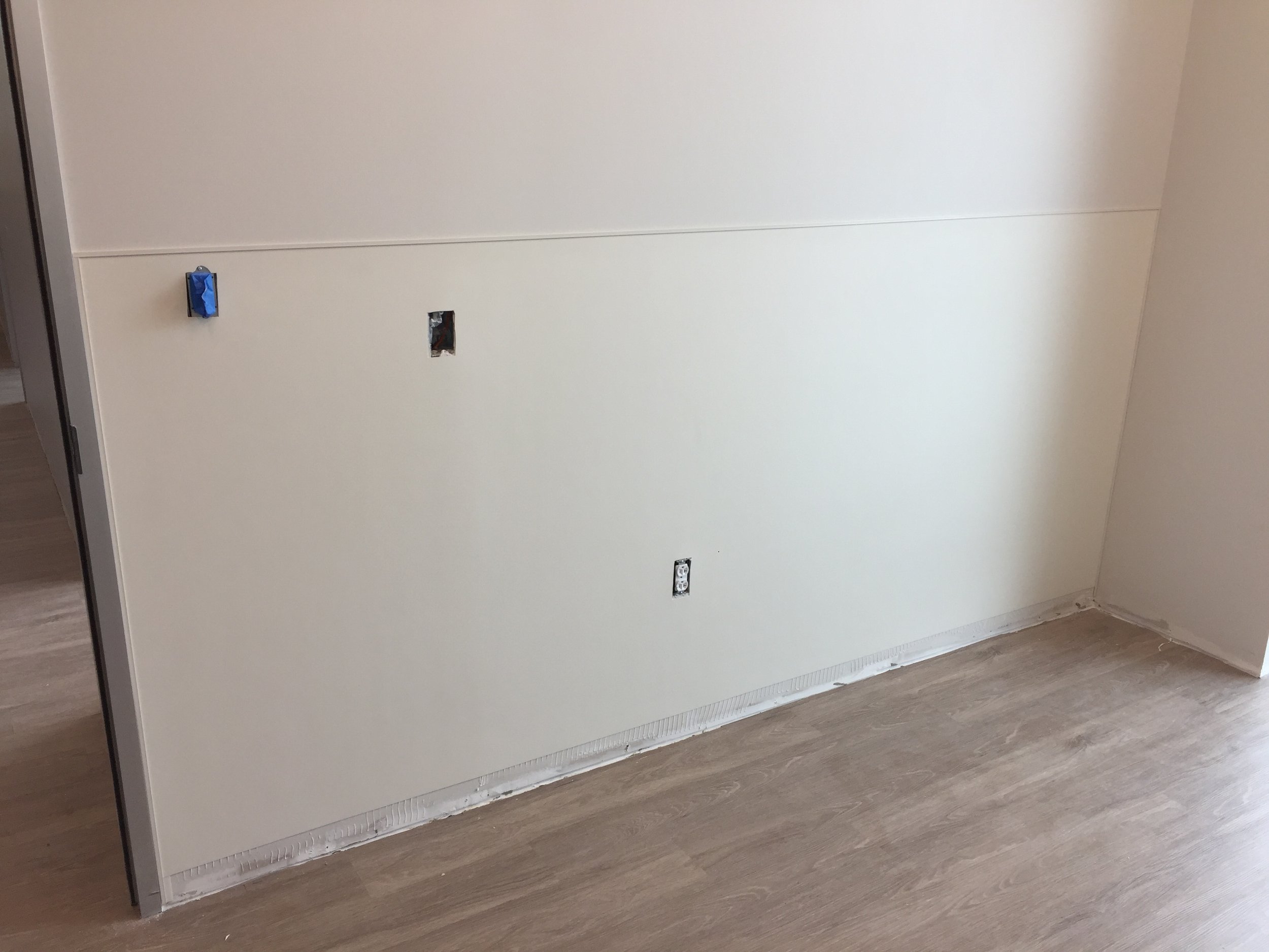 A partially finished wall with two electrical outlets and a hole where a switch may be installed, with some electrical wiring visible. The wall has a cable running along the middle, and a baseboard heater at the bottom.