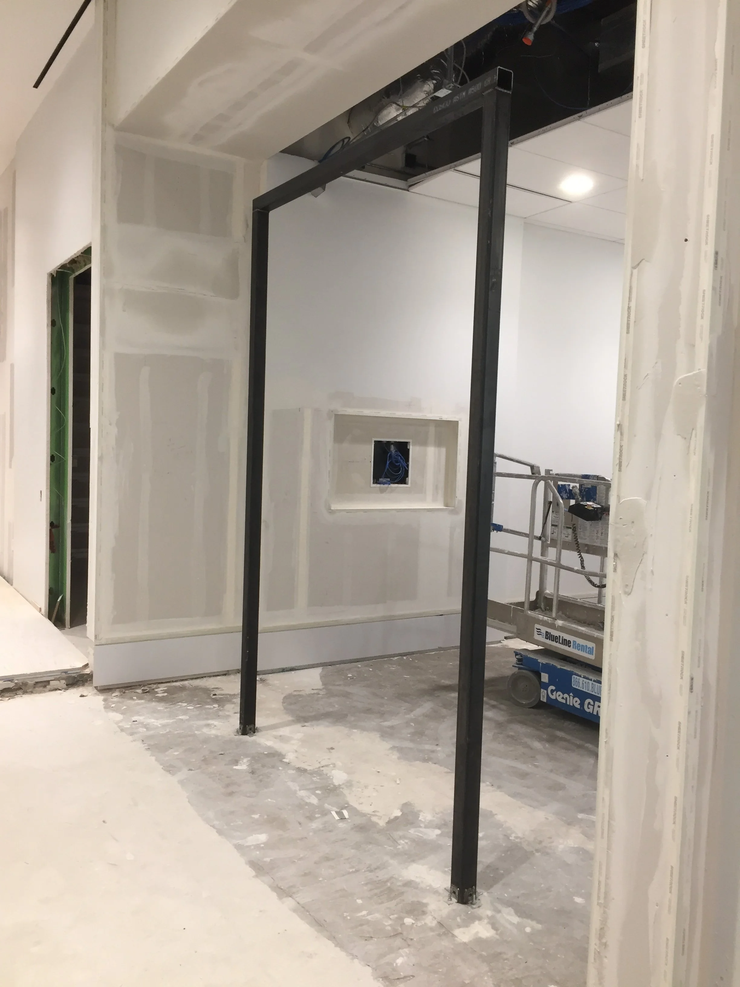 Interior of a building under construction with drywall walls, exposed wiring, and a metal support structure in the center of the room.