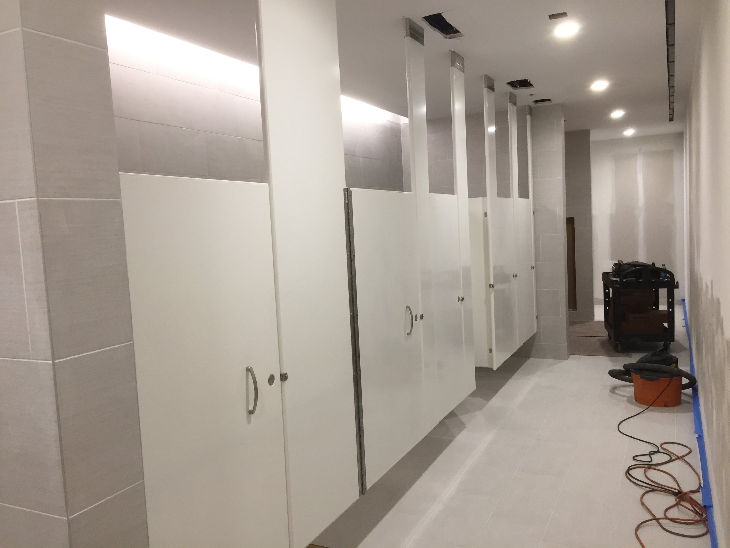 The image shows a partially constructed or renovated public restroom with multiple white stall doors, some of which are missing handles or locks. The ceiling has open spaces with exposed vents, and the floor tiles are light-colored. There are constru