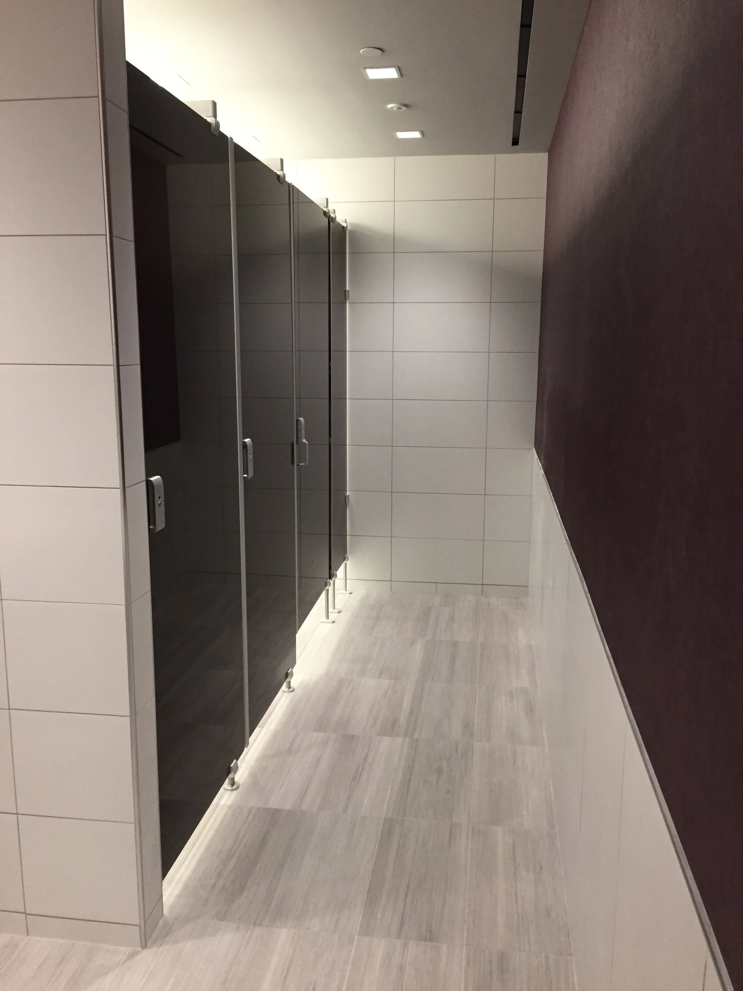 A restroom with four vacant stalls with glass doors, a tiled bathroom wall, light grey flooring, and a dark brown counter on the right side.