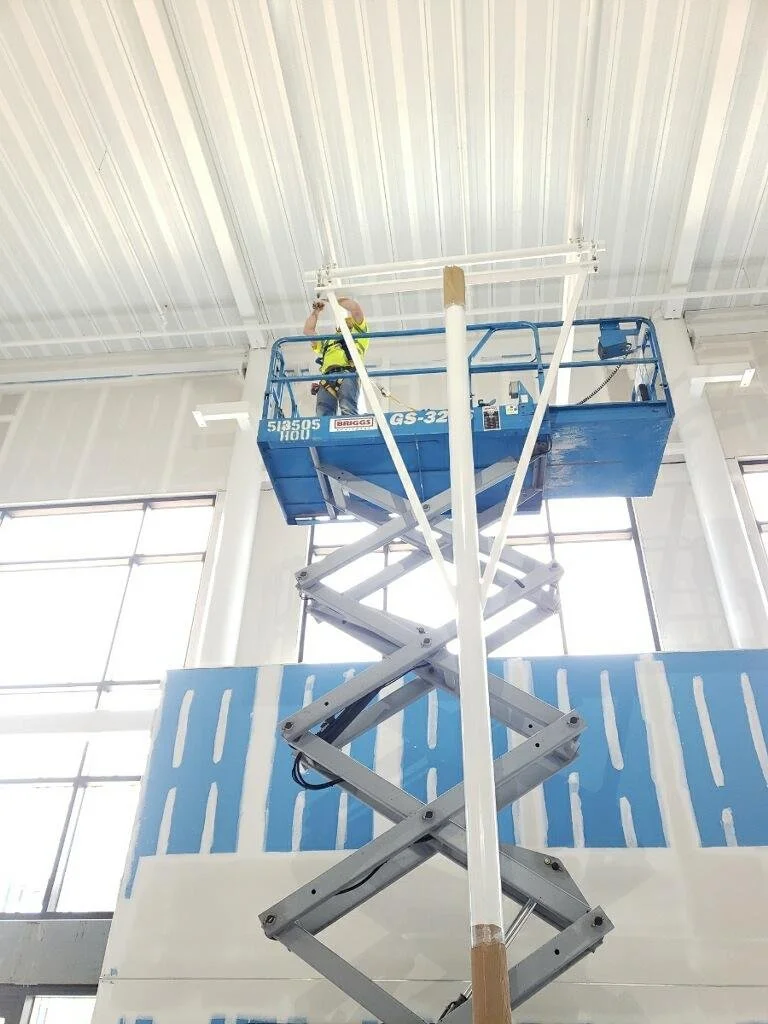 A worker in a yellow safety vest on a blue scissor lift working on the ceiling of a large industrial or commercial space.