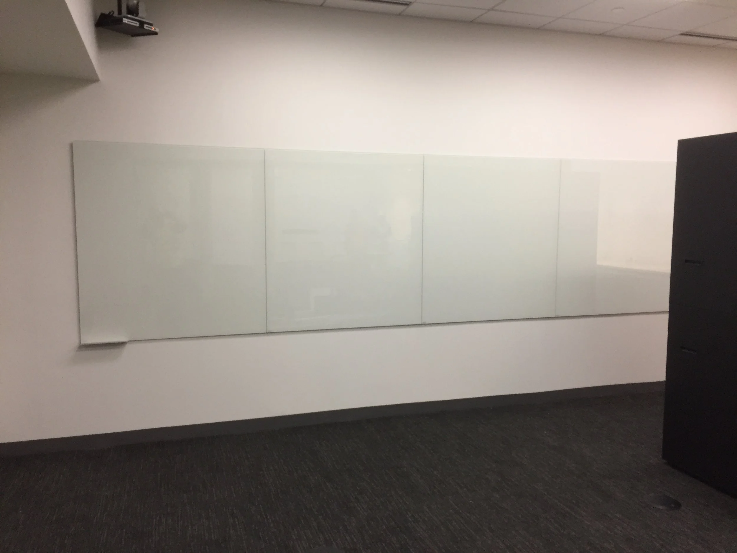 A plain white wall with a black object mounted near the ceiling, a whiteboard, and a dark carpeted floor.