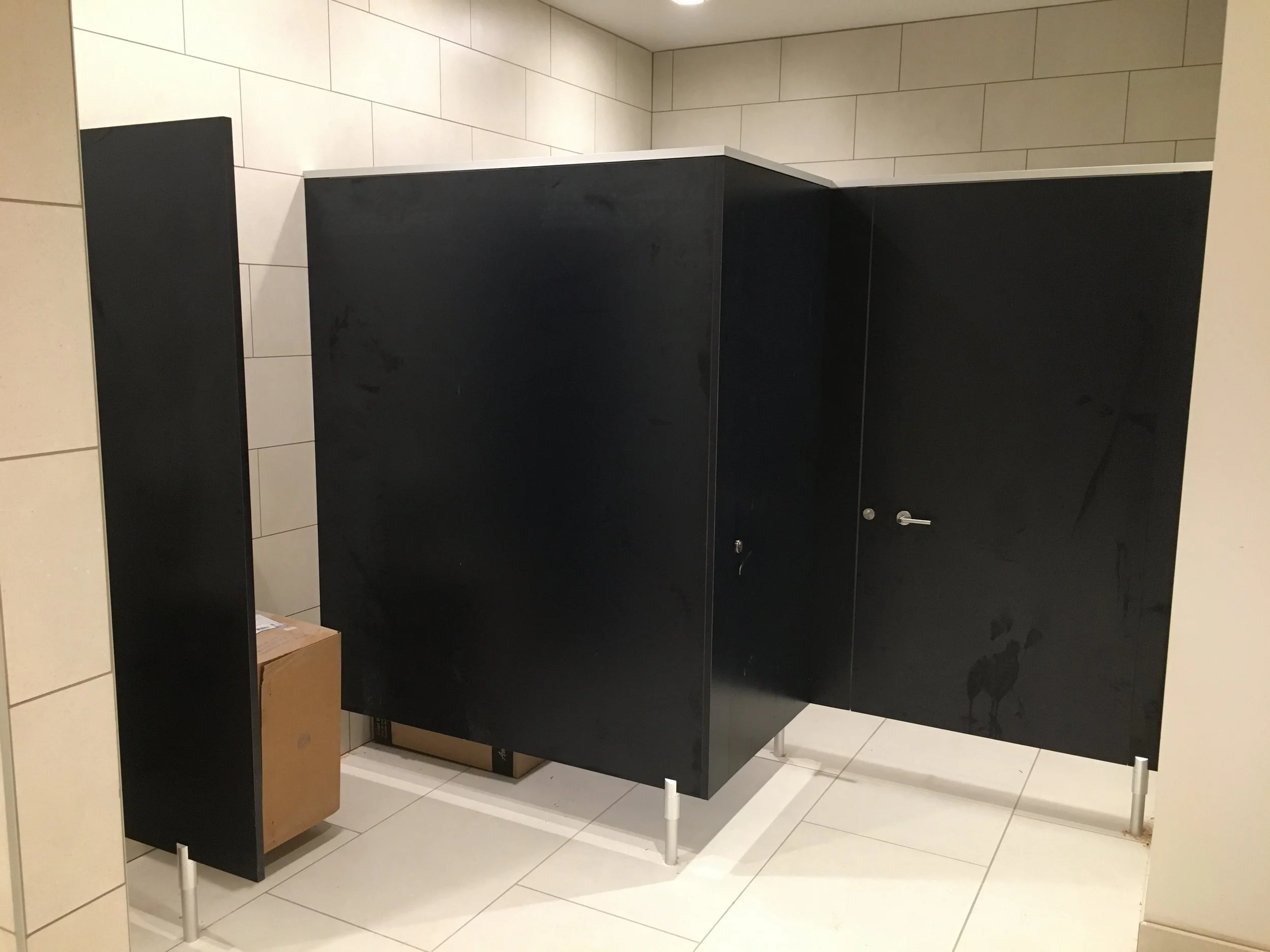A restroom stall with black doors and white top edges, set against beige tiled walls and floor, with a cardboard box and a few white poles in the foreground.