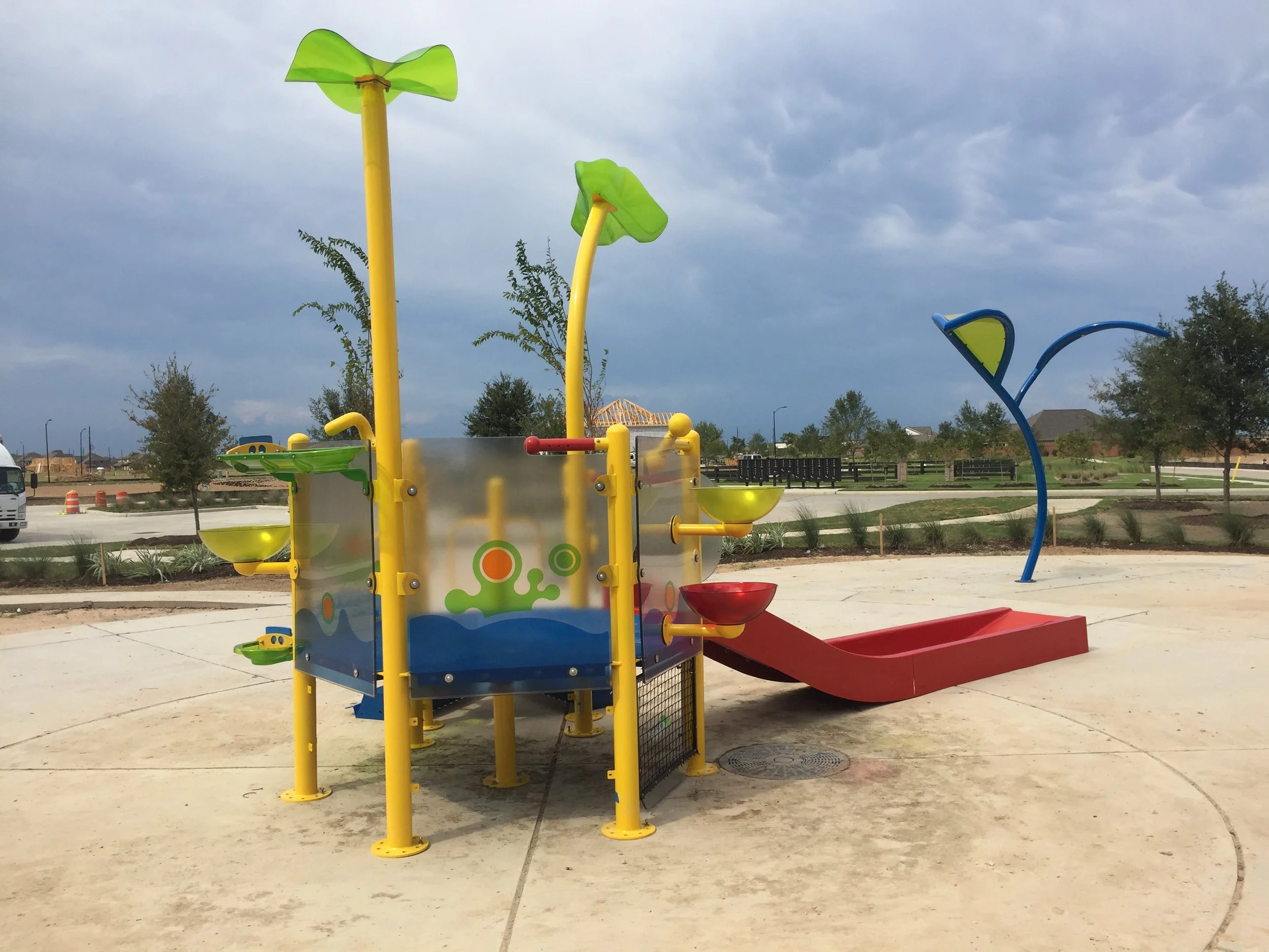 Colorful playground equipment with yellow, green, and blue components, including a slide and interactive play panels, set in an outdoor park area with trees, a fence, and overcast sky in the background.