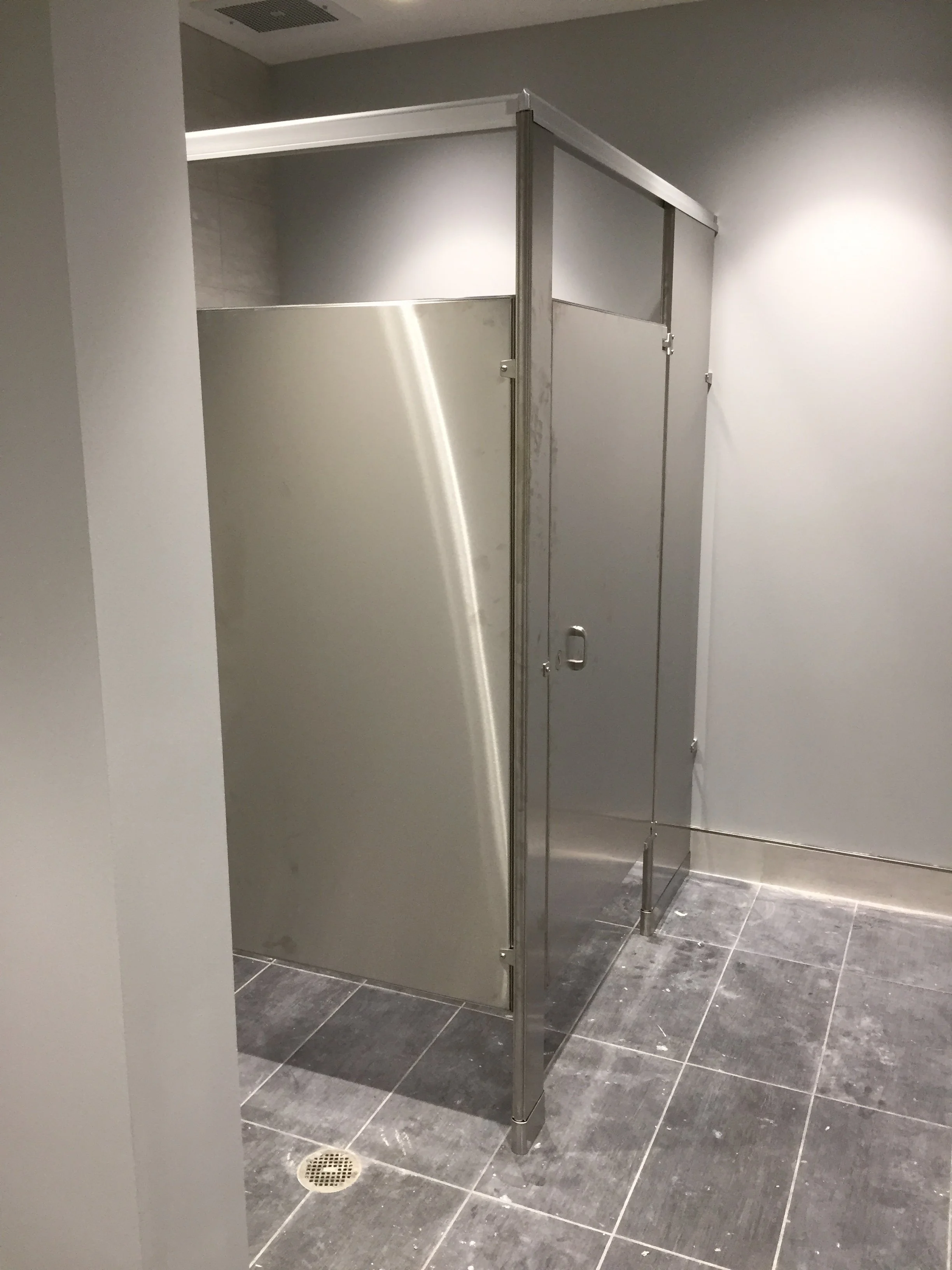 A partially enclosed public restroom stall with gray metal doors and framing, gray tiled floor, and a floor drain visible.
