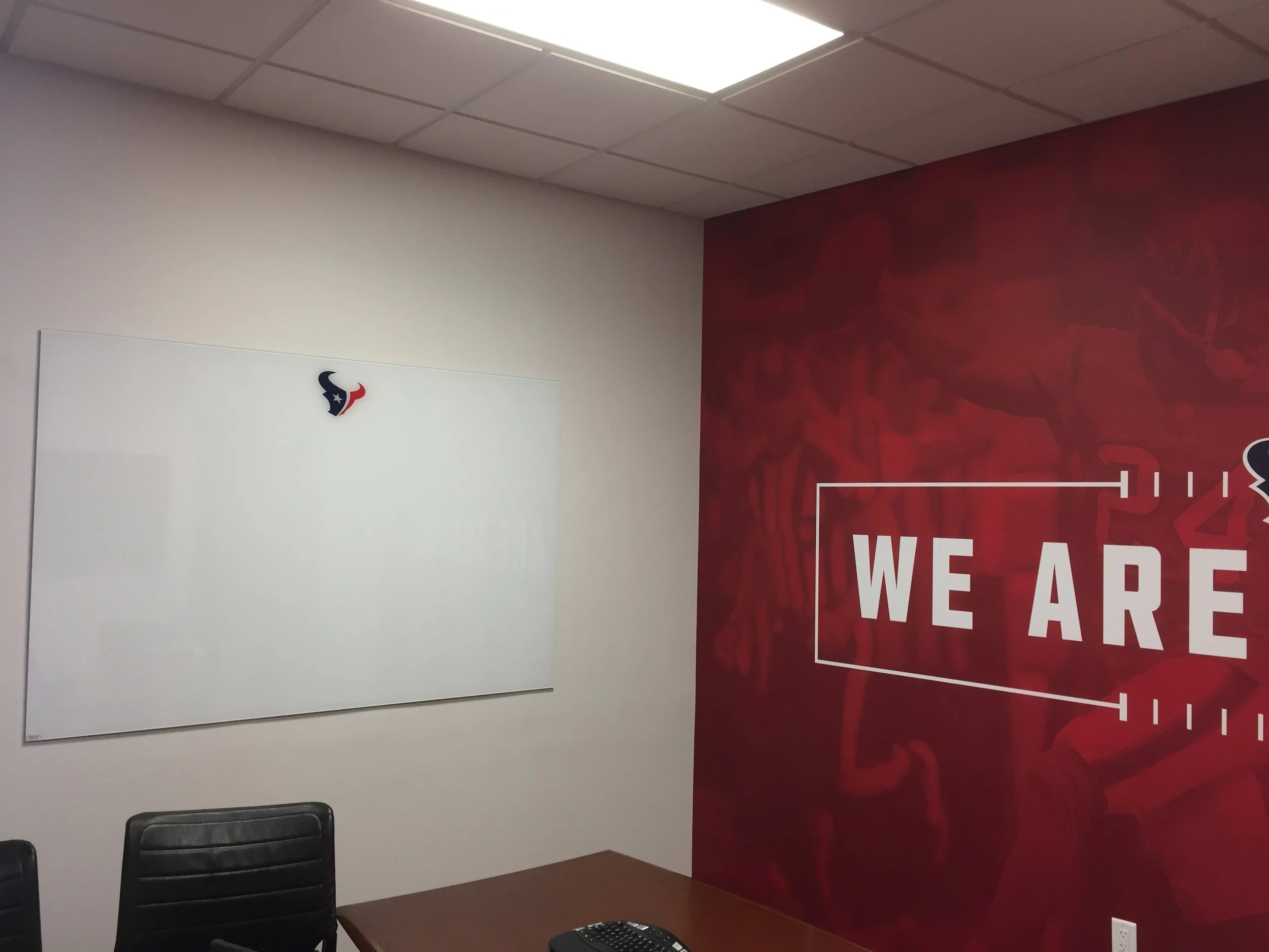 Interior of a conference room with a Houston Texans logo on the whiteboard, black chairs, and a red wall with the words "WE ARE" partially visible.