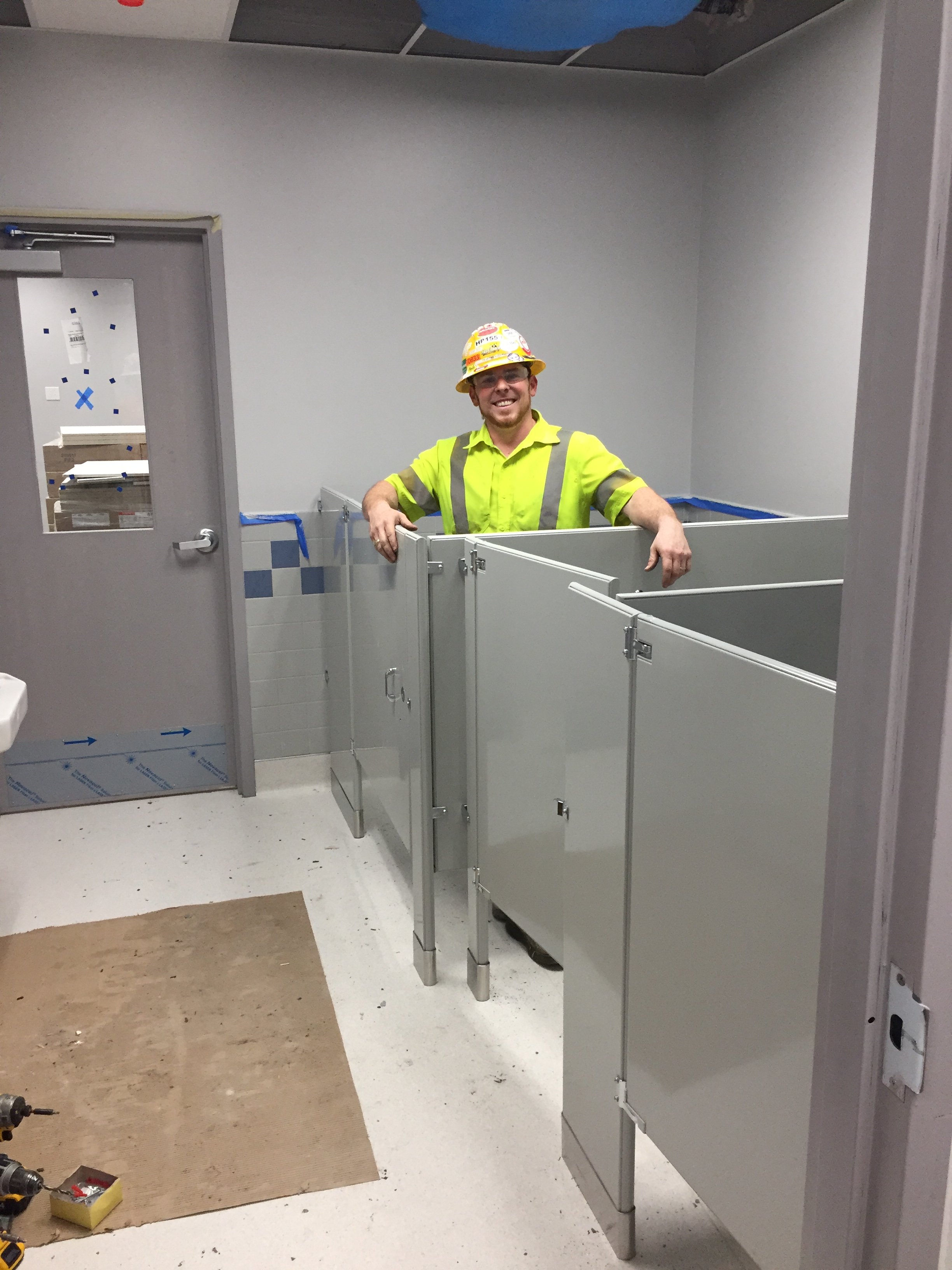 Construction worker wearing a yellow safety vest and yellow hard hat smiling inside a bathroom stall under construction, with bathroom stalls partially installed and tools on the floor.