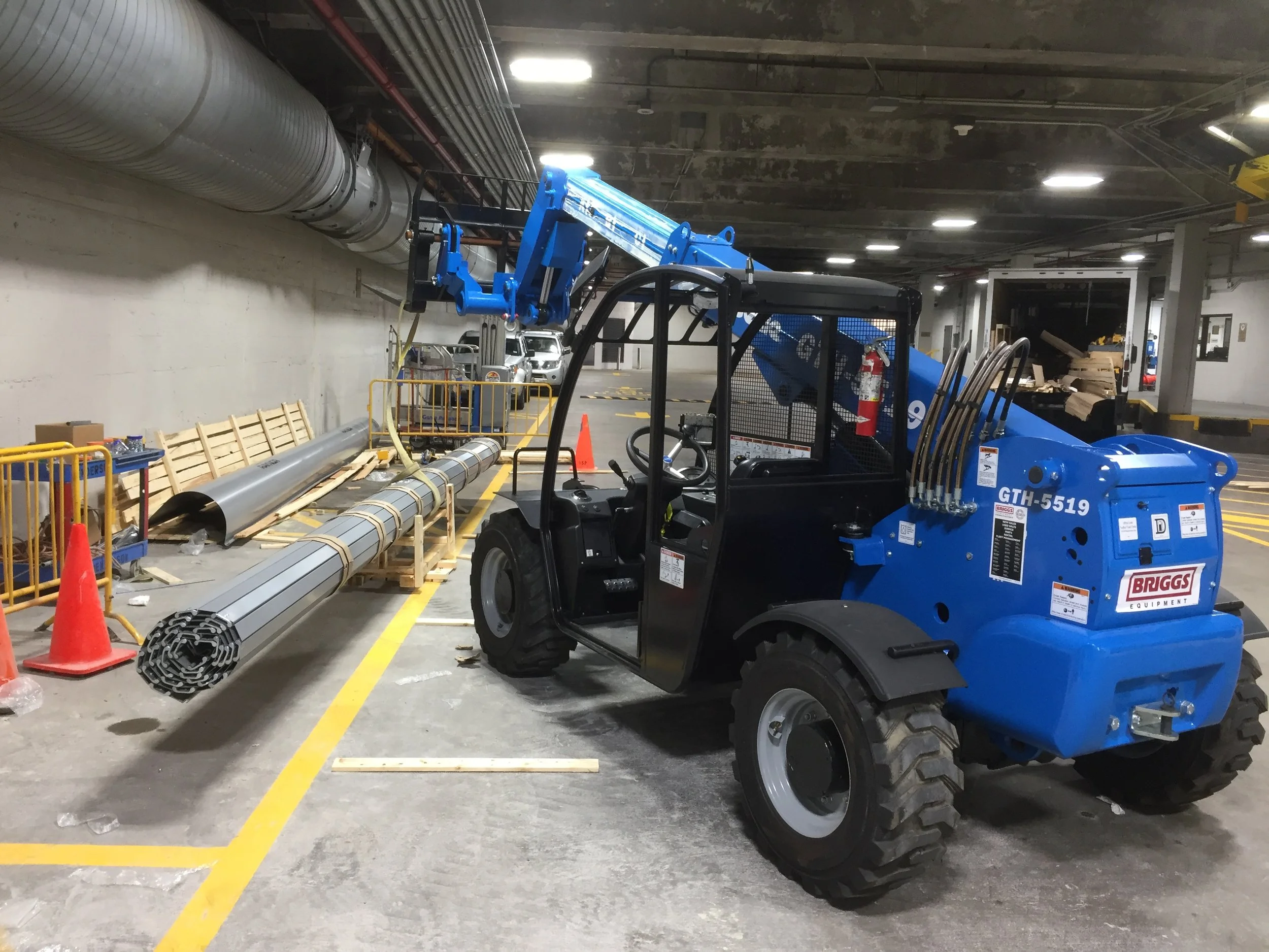 A blue forklift lifting a large pipe in an underground parking garage or storage area.