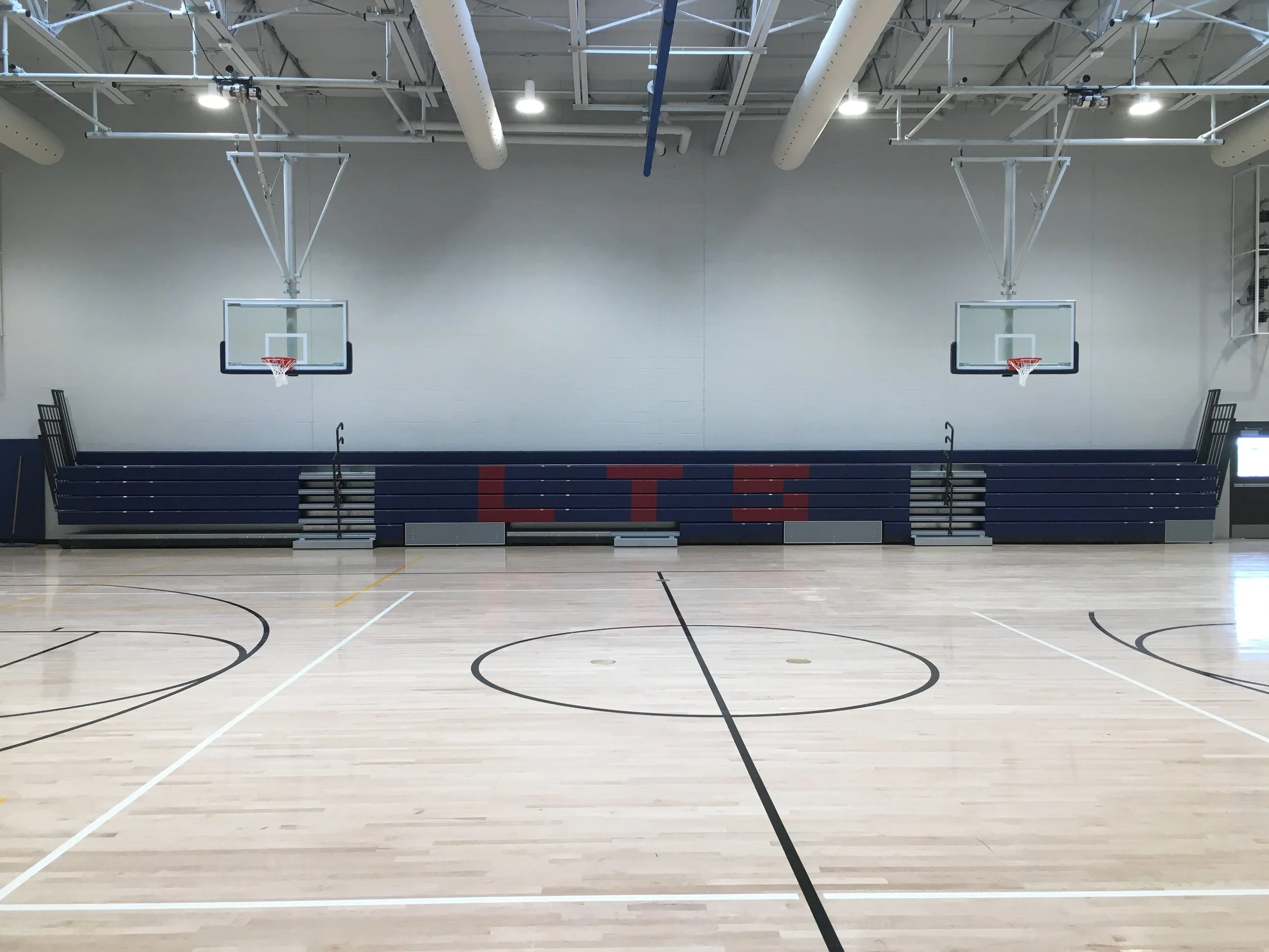 Indoor basketball gymnasium with two basketball hoops, grey walls, and bleachers at the side.